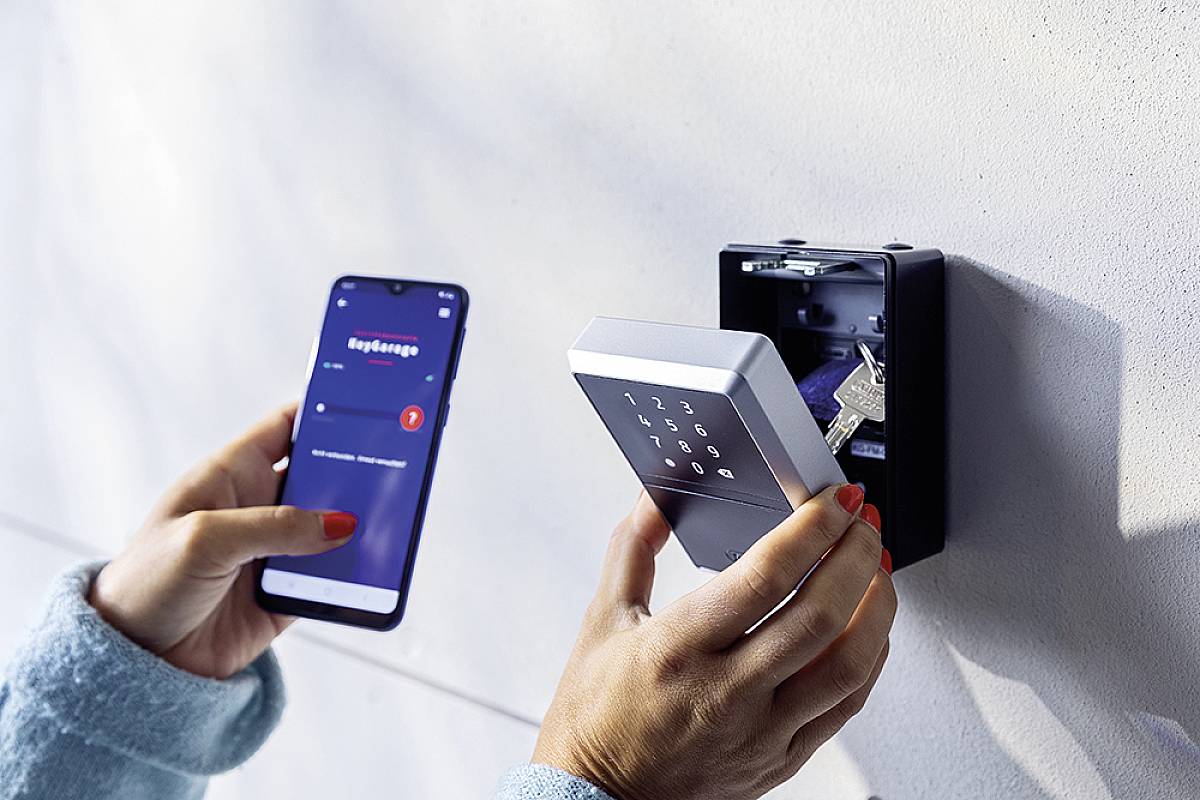 En person holder en smartphone med en åben app i venstre hånd og en elektronisk nøgleboks i højre hånd.