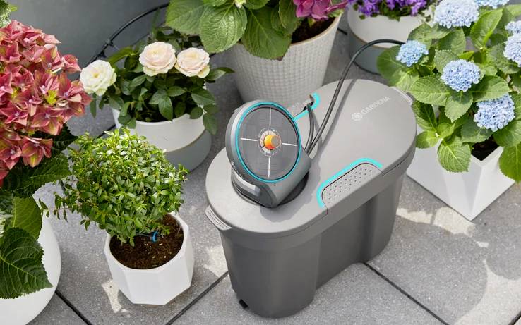 Flere blomsterkrukker med farverige blomster på en terrasse, ved siden af et grå-sort vandingssystem med slanger.