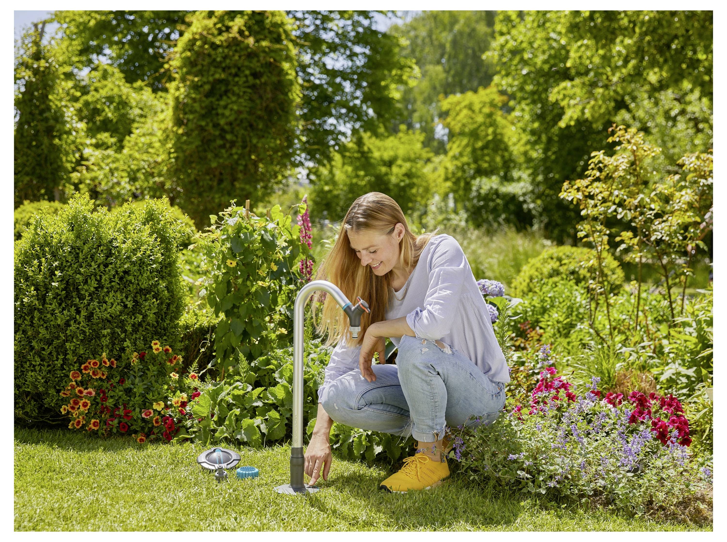 En kvinde knæler på græsset i en have og tænder for en vandhane for at vande planter omgivet af levende blomster og grønt under en solskinsdag.