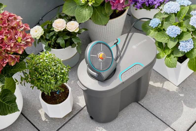 En havevandingsanordning er placeret mellem potteplanter med blomster på en flisebelagt terrasse, hvilket viser dens anvendelse til automatisk plantevanding.