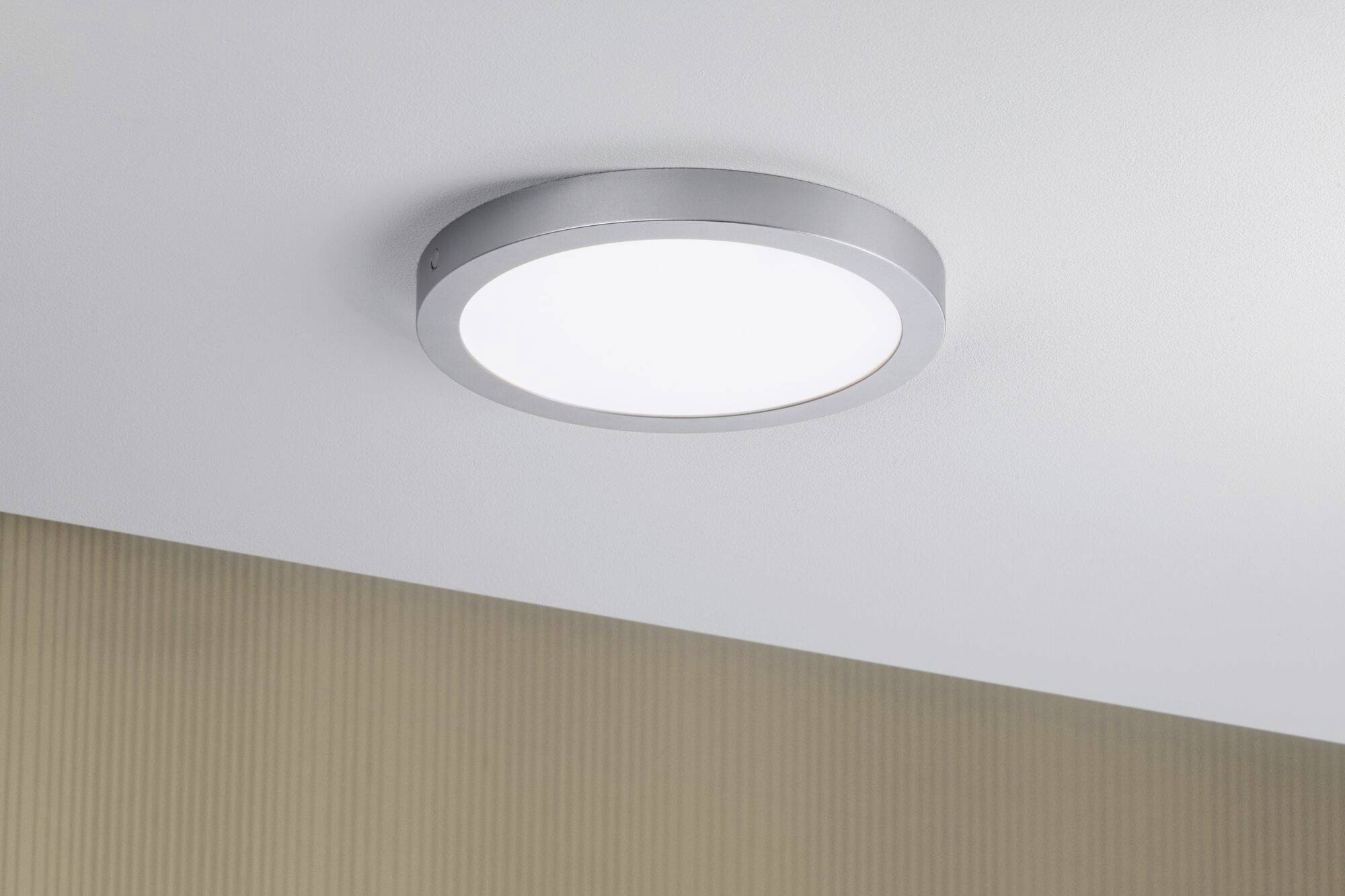 Rund loftlampe med sølvkant monteret på hvid loft, som lyser kraftigt op. Nedenunder en væg med diskret tekstur.
