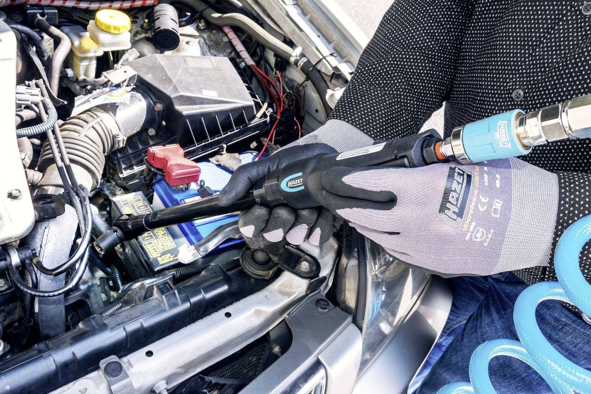 En person kontrollerer bilens motor med et måleapparat, mens vedkommende bærer beskyttelseshandsker. Motorrummet er detaljeret synligt.