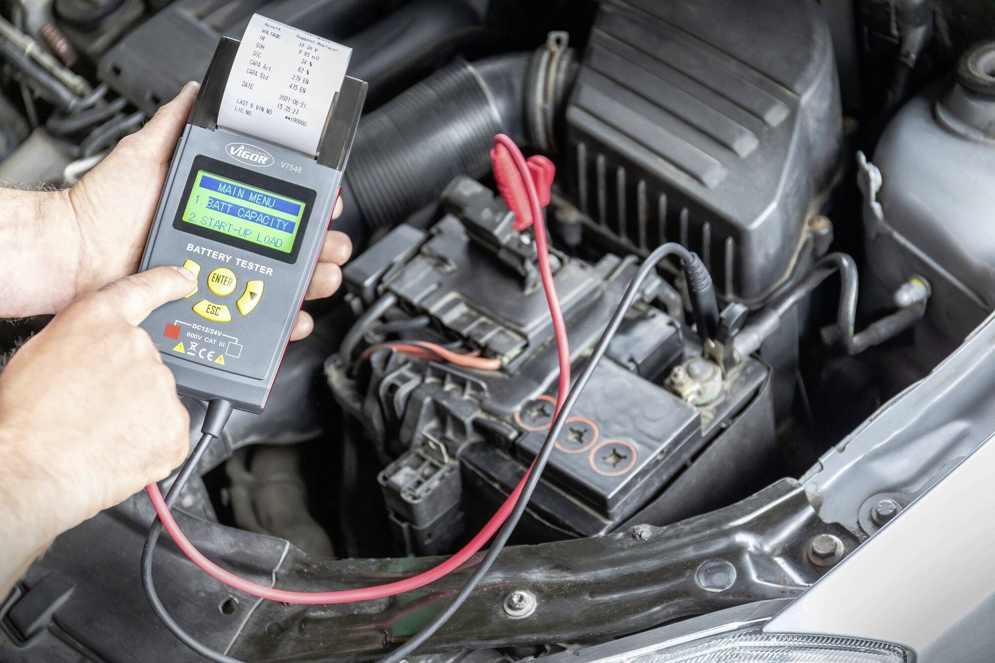 En person kontrollerer bilbatteriet med en digital tester i motorrummet. Enheden viser måleværdier på skærmen, mens kabler er tilsluttet batteriet.