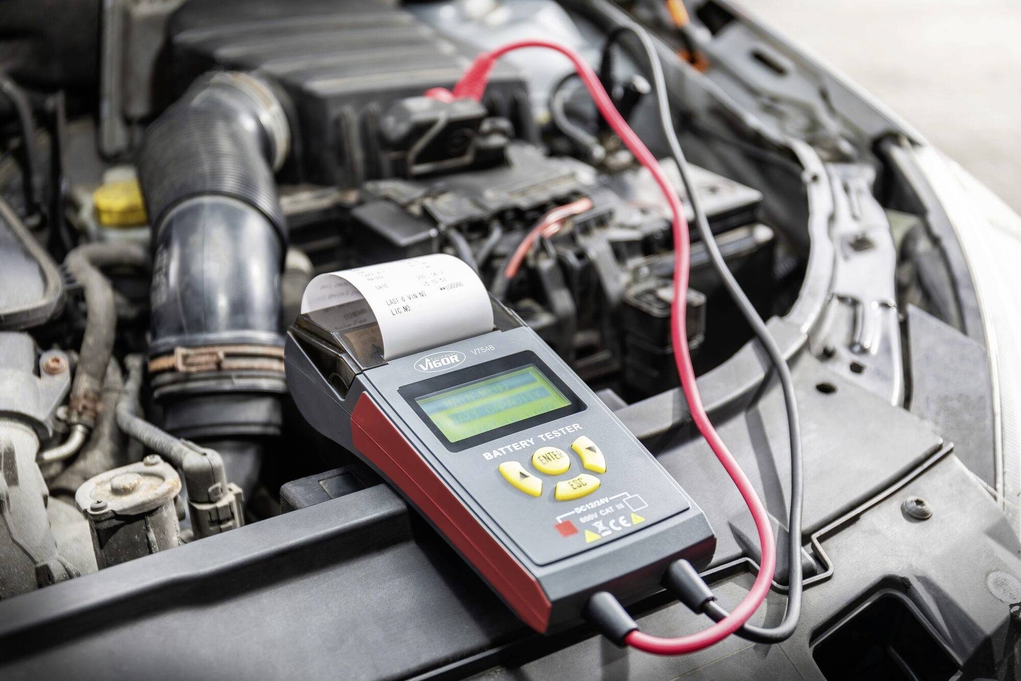 En batteritester er tilsluttet bilbatteriet for at kontrollere opladningstilstanden. Displayet viser et resultat på en lille skærm.