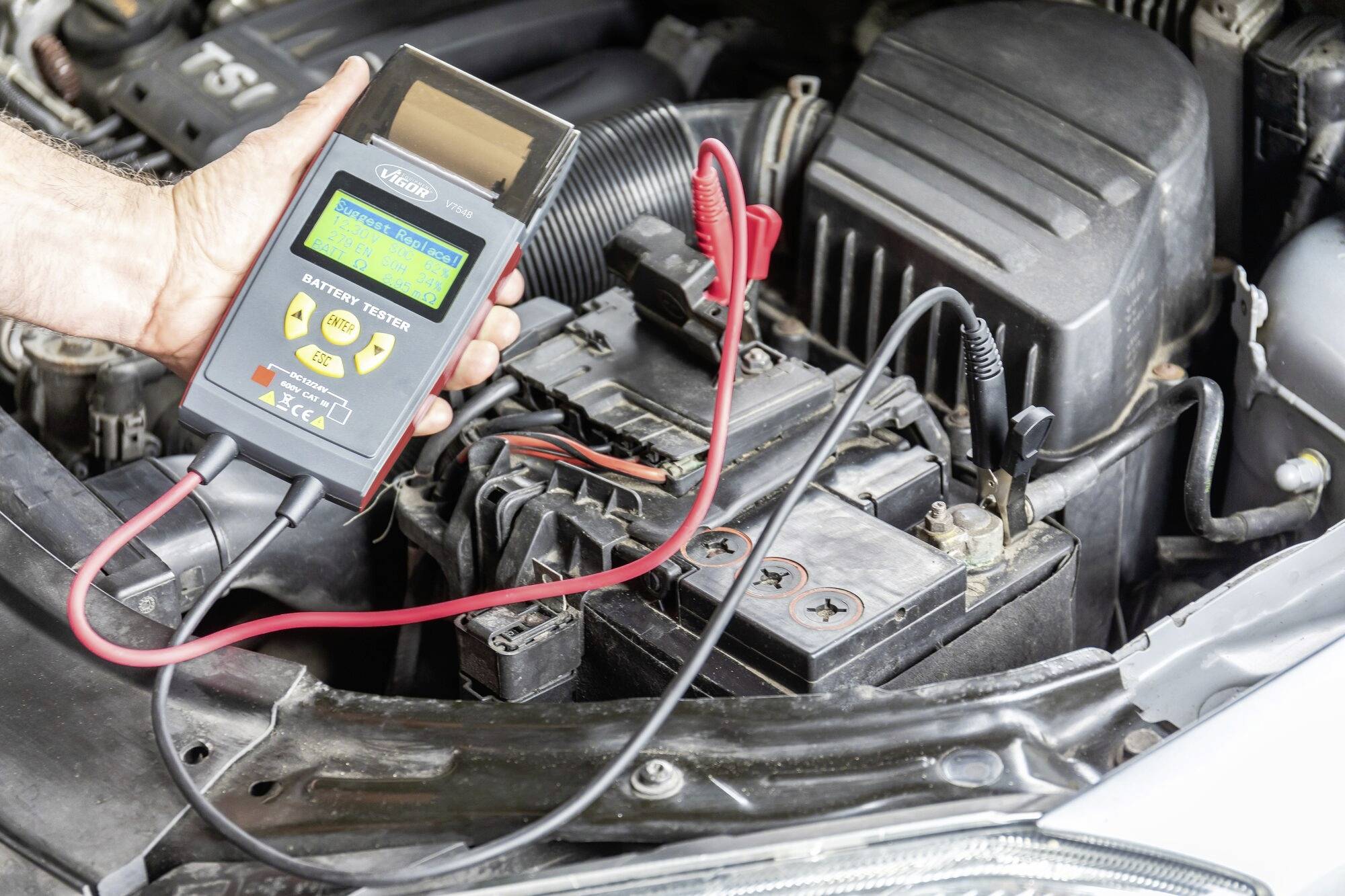 En person holder en batteritester ved den åbne motorhjelm. Enheden viser oplysninger om batteriets tilstand.