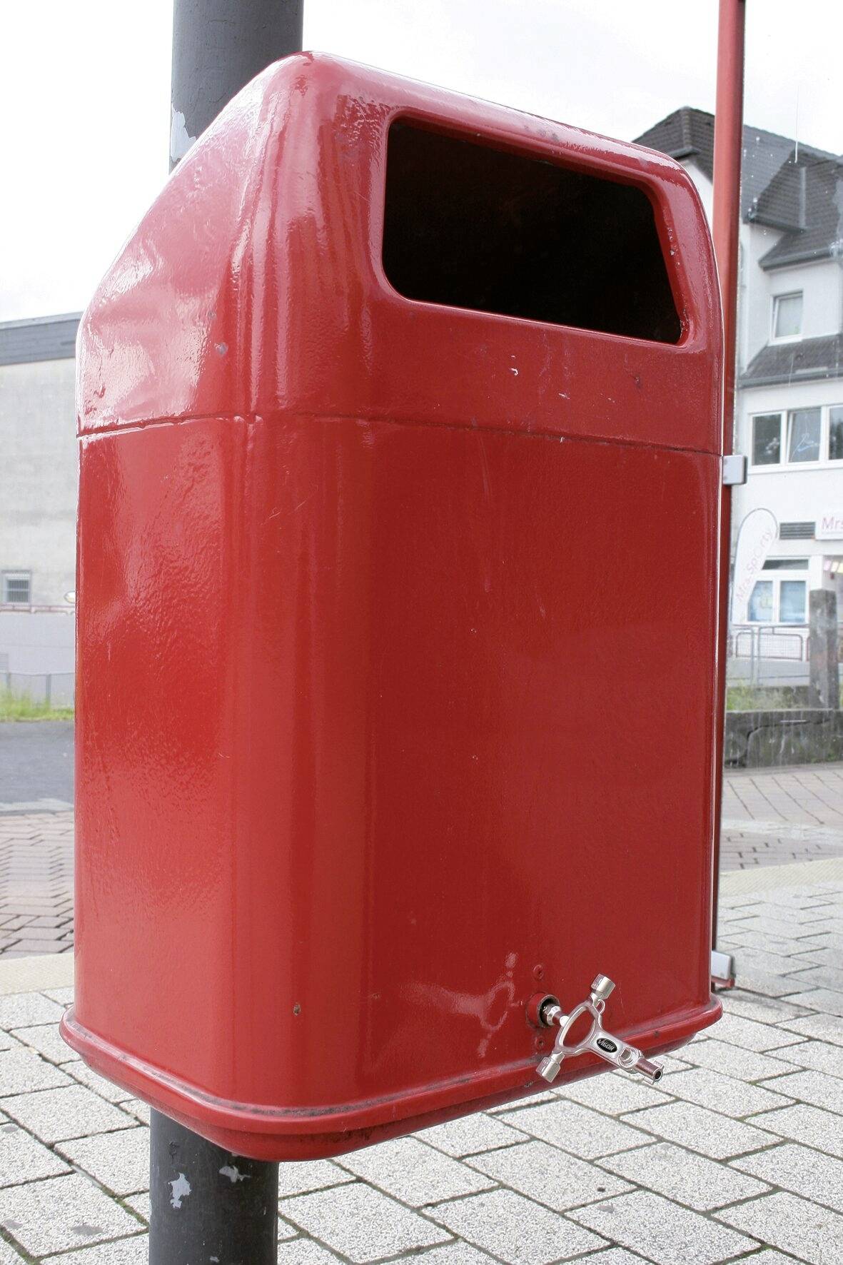 Rød skraldespand ved en vej, monteret på en metalstang. I baggrunden ses bygninger.