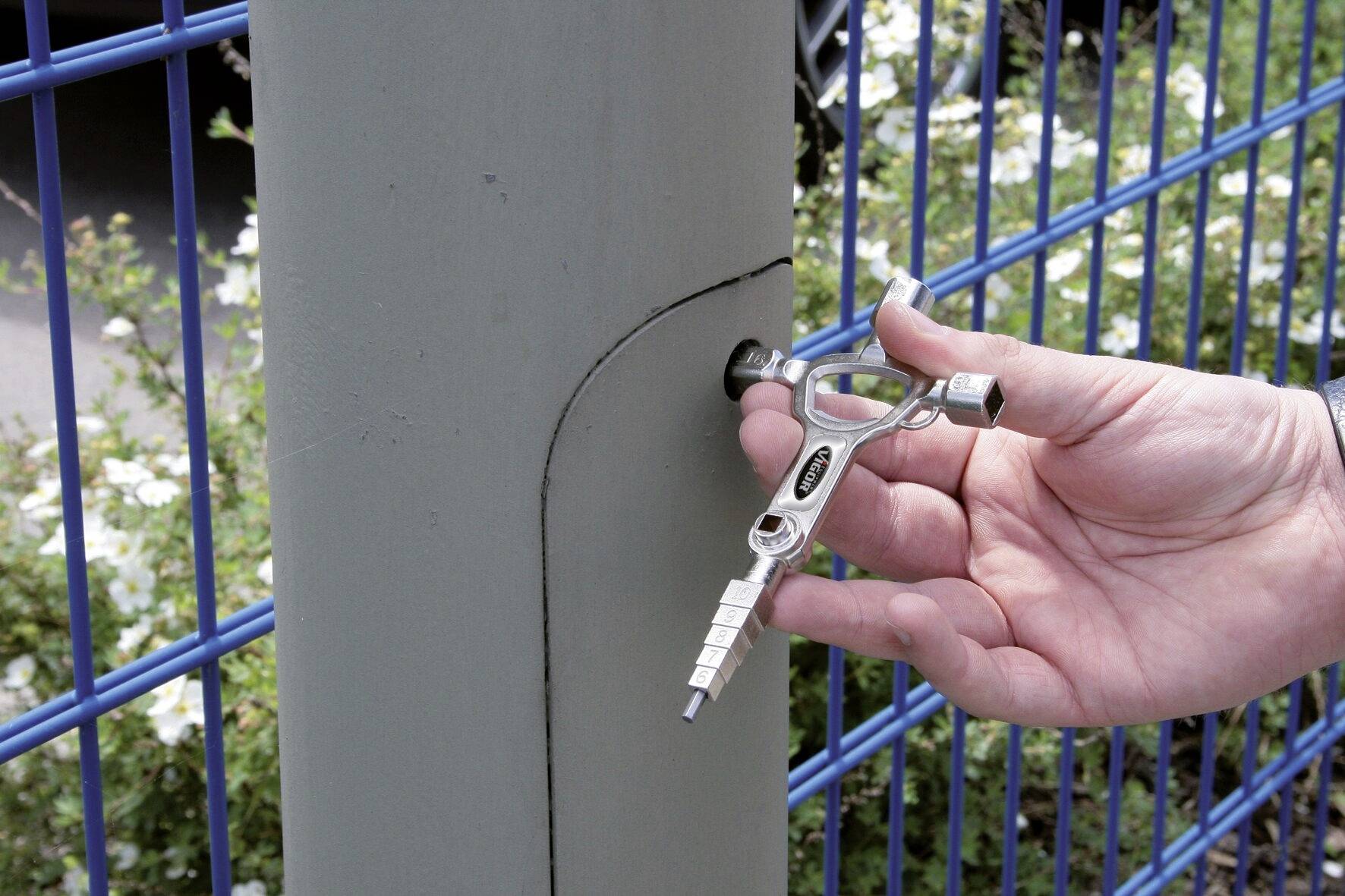 En hånd bruger en firkantet nøgle til at åbne en lås på en metalpæl ved siden af et blåt trådhegn.