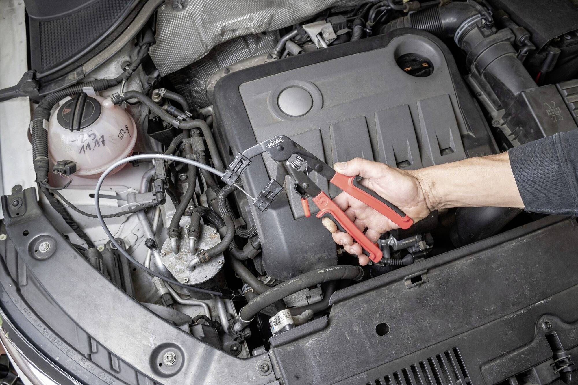 En hånd holder en tang over en åben motorrum i en bil og viser brugen af værktøjet ved vedligeholdelse af køretøjet.