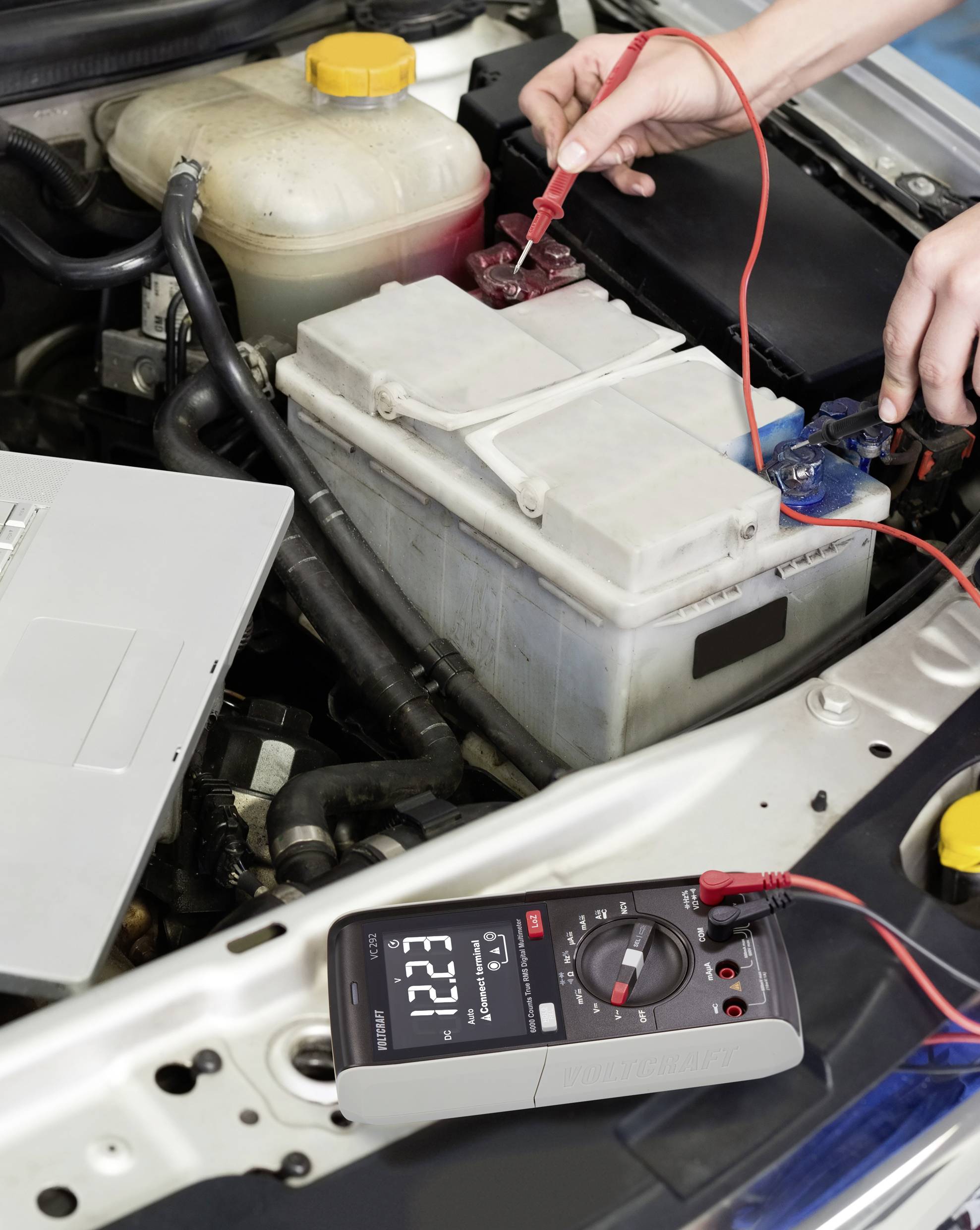 En person måler spændingen på en bilbatteri i motorrummet ved hjælp af et multimeter. Displayet viser '12,21'.
