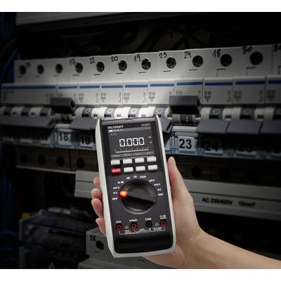 En hånd holder et digitalt multimeter foran en åben sikringsskabe med flere sikringsautomater.