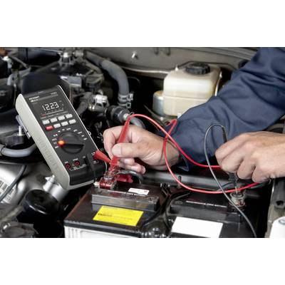 En person måler spændingen på en bilbatteri i motorrummet ved hjælp af et digitalt multimeter. Fokus på hænderne og måleapparatet.