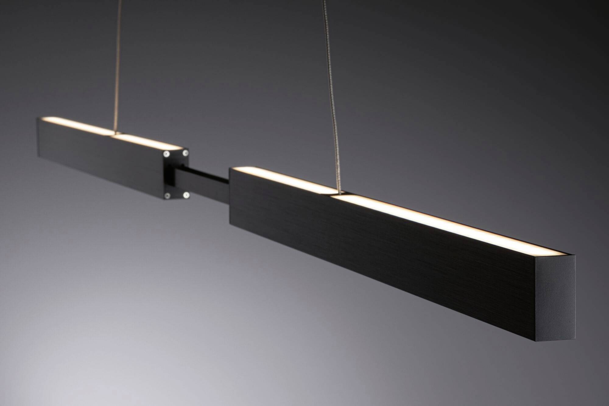 En moderne, aflang loftslampe i sort metal svæver i et mørkt rum. Den har to rektangulære lysbjælker.