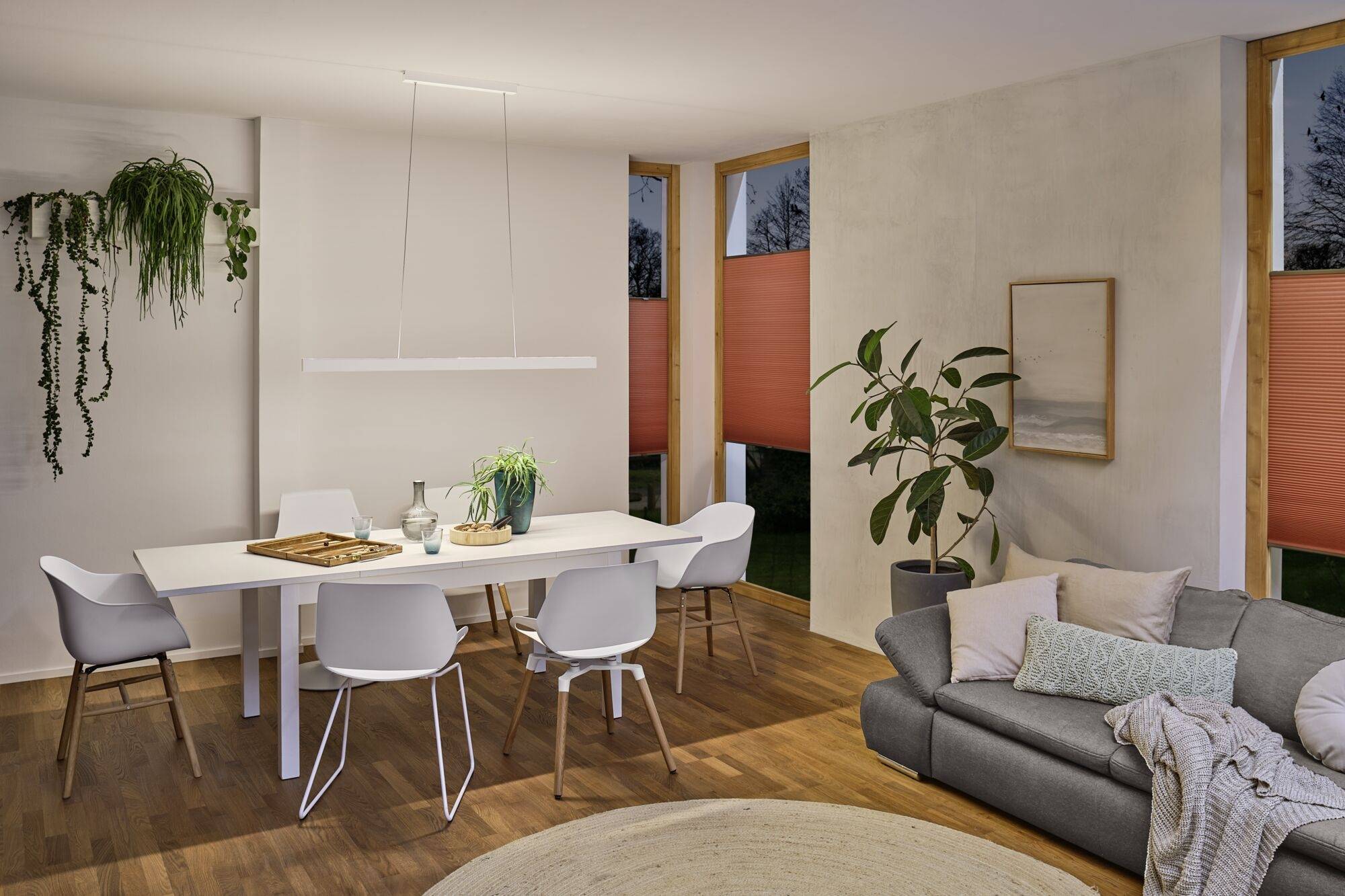 Spisestue med et moderne hvidt bord, seks stole, grå sofa, planter og et rektangulært kunstværk, varmt belyst.
