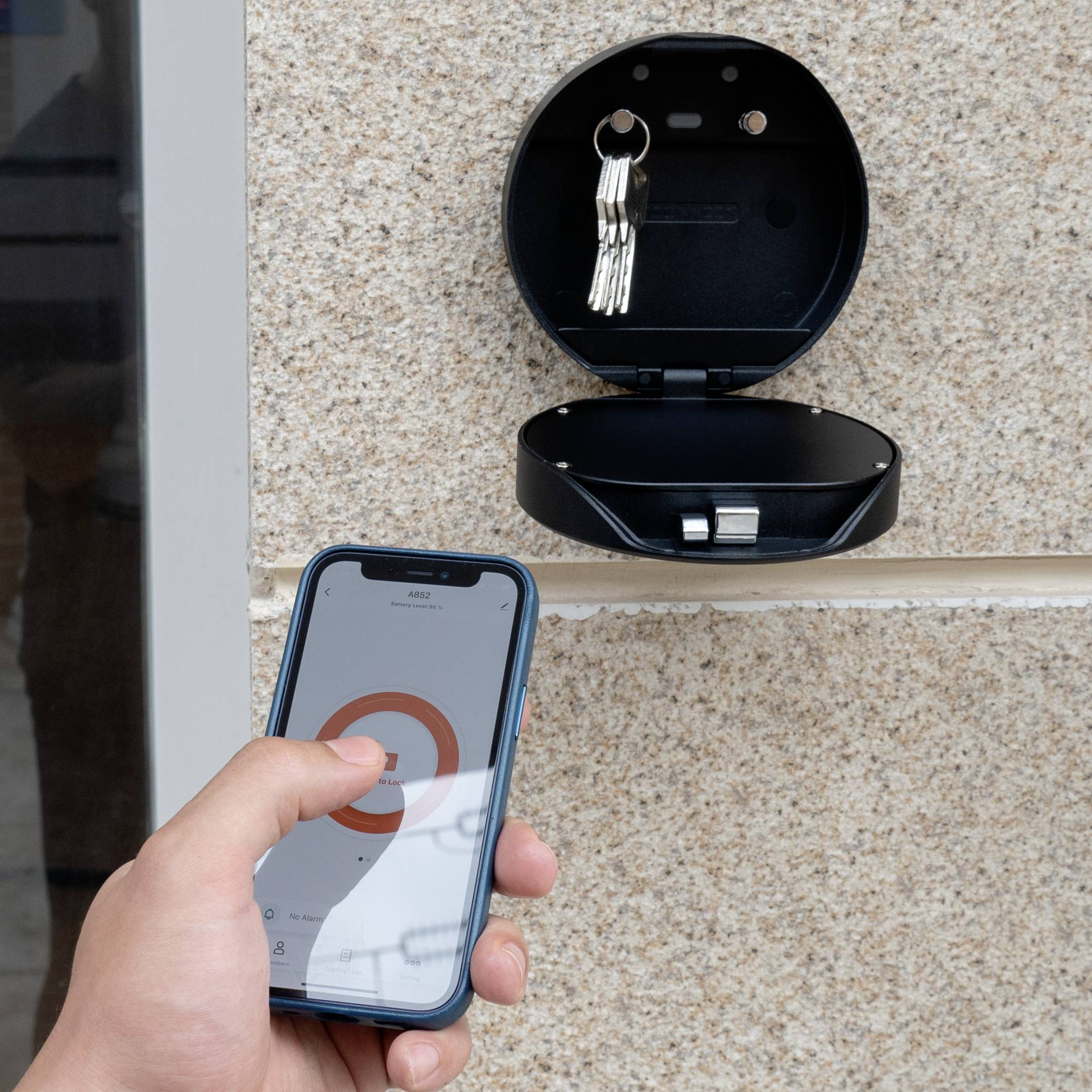 En hånd holder en smartphone med en app. Et vægbeslag med et åbent, rundt hylster viser de opbevarede nøgler.