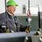 En arbejder med en grøn hjelm betjener en kran i en industribygning. Metalplader bliver flyttet. Sikkerhedssko og handsker er synlige.