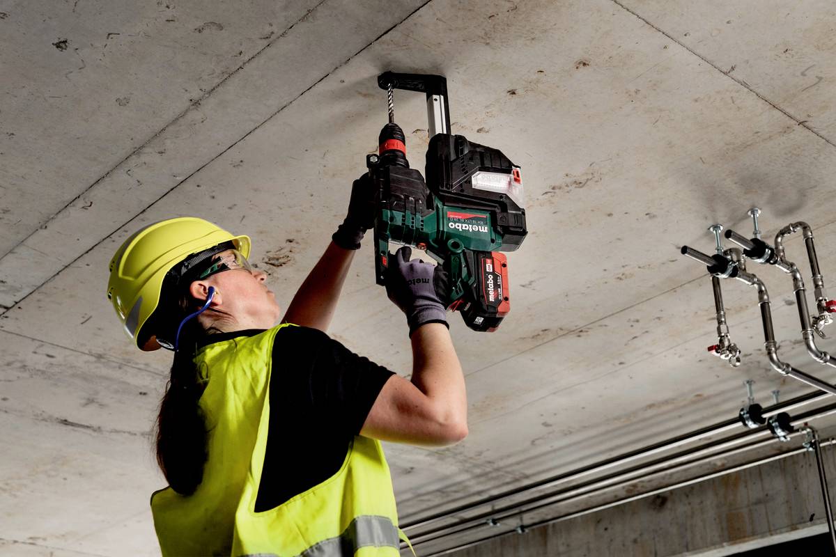 En person med sikkerhedshjelm og refleksvest borer i et loft med et elværktøj i et industrielt miljø.