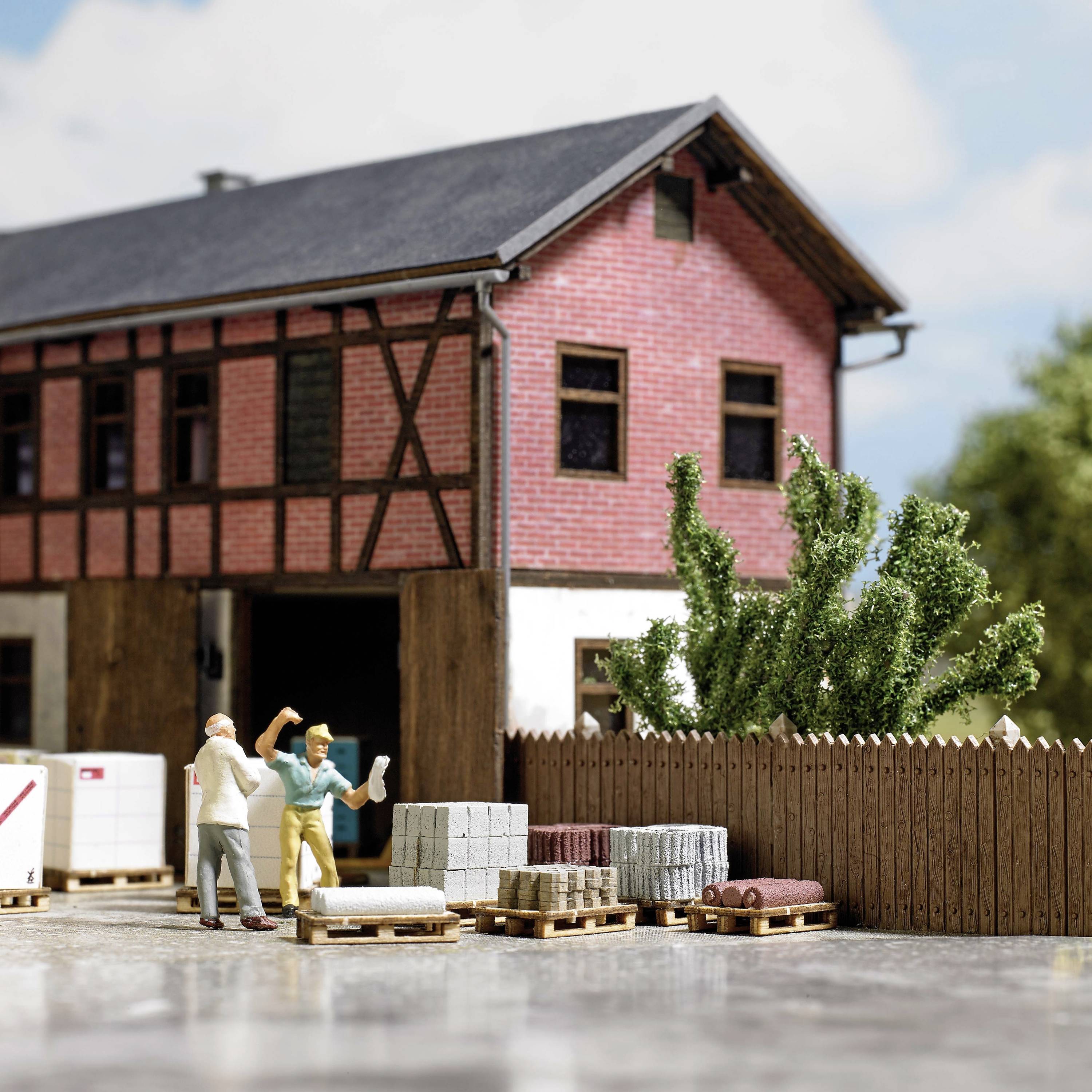 To miniaturfigurer i arbejdstøj står foran en model af en fabrik med teglsten og trælammer, omgivet af paller og planter.
