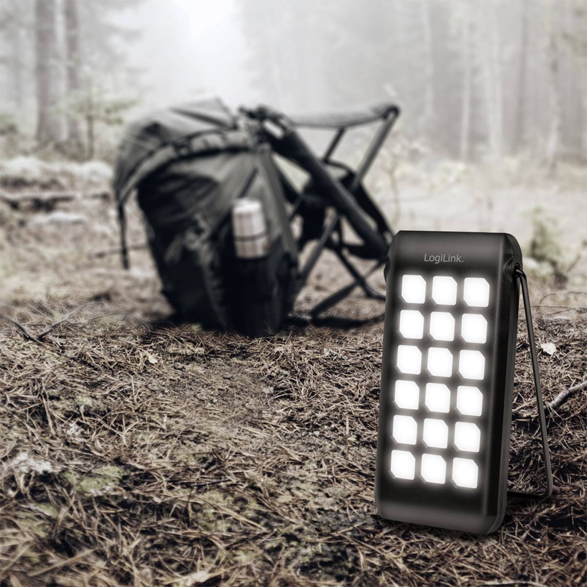 Bærbar LED-lampe i forgrunden, placeret i skoven; i baggrunden en rygsæk og en campingstol, som indikerer en udflugt i naturen.