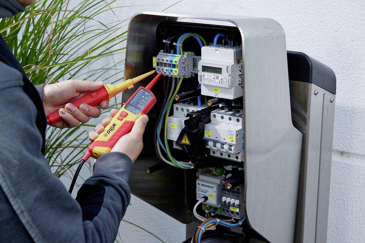 En tekniker kontrollerer en elektrisk installation med et multimeter. Flere kabler og sikringer er synlige.