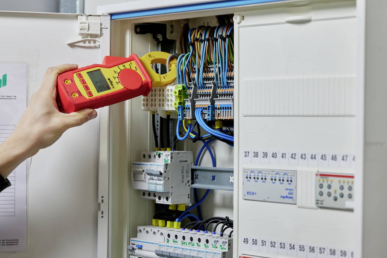 En person måler elektrisk spænding i en åben tavle ved hjælp af et multimeter. Talrige farverige kabler er synlige.