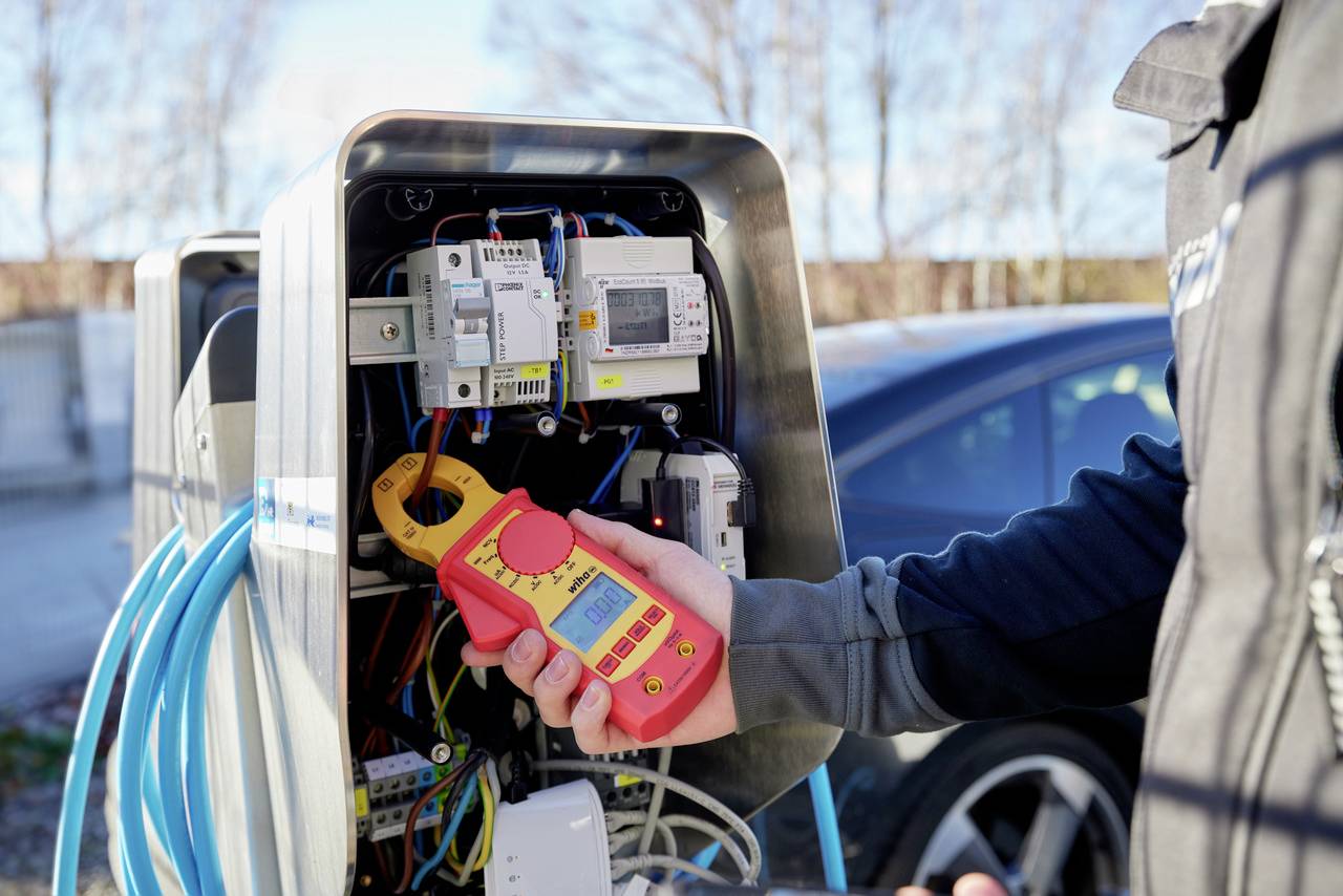 En person holder en måleenhed ved siden af en åben ladestander til elbiler og kontrollerer den elektriske forbindelse.