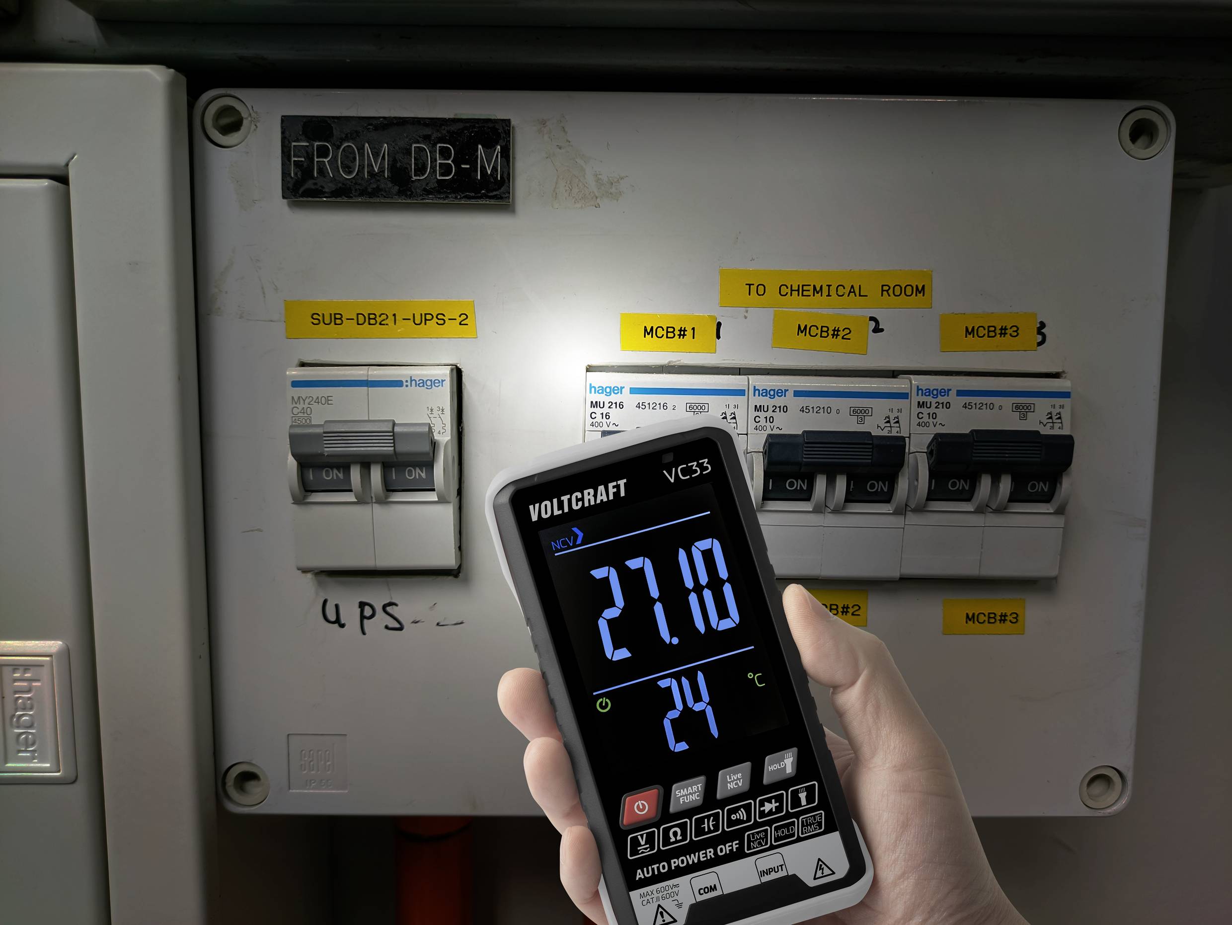 En hånd holder et digitalt voltmeter foran en sikringstavle. Skærmen viser 271,0 volt og 24 grader celsius.