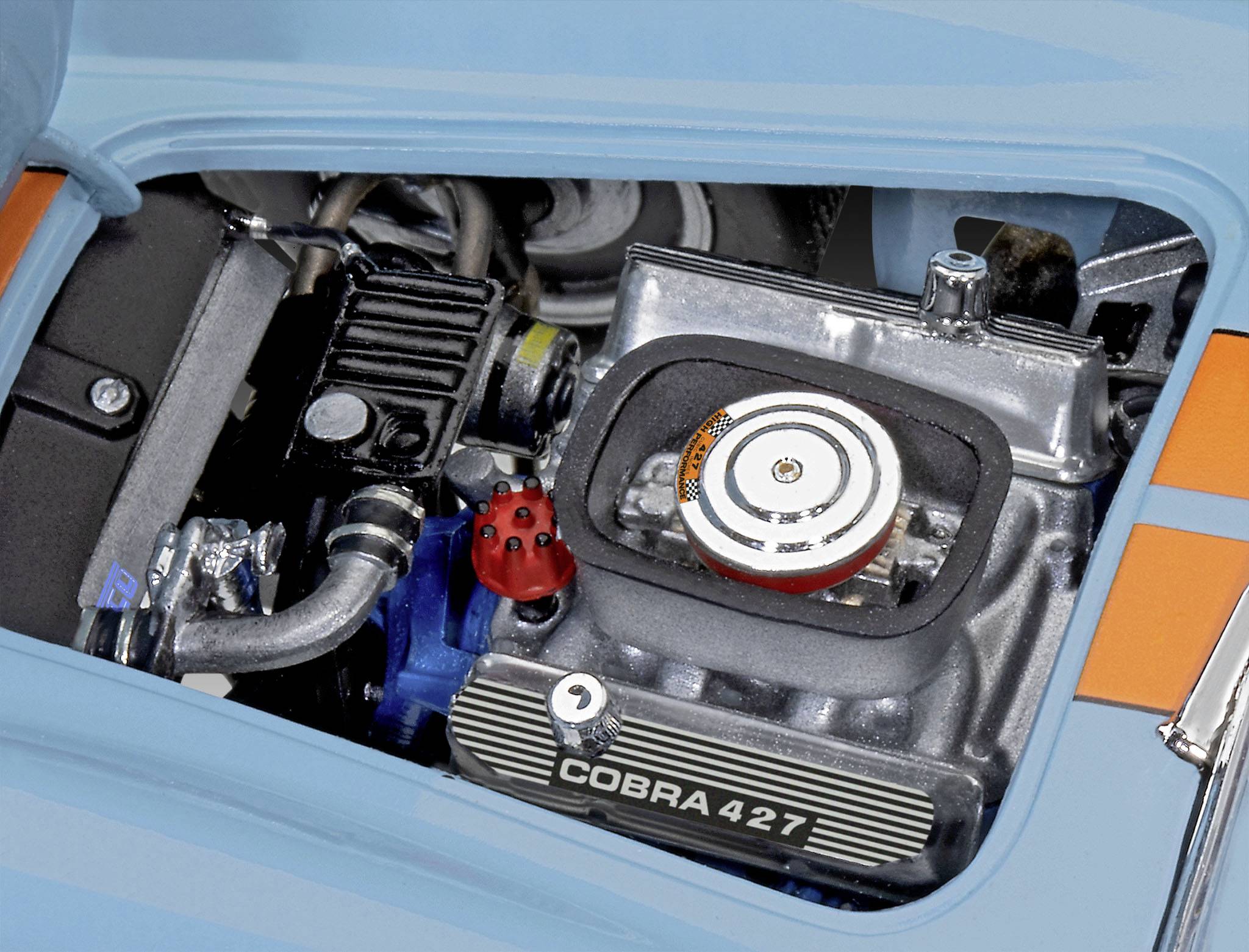 Et åbent motorrum på en blå bil viser en Cobra 427-motor i nærbillede, som er sølvfarvet med metaldetaljer.