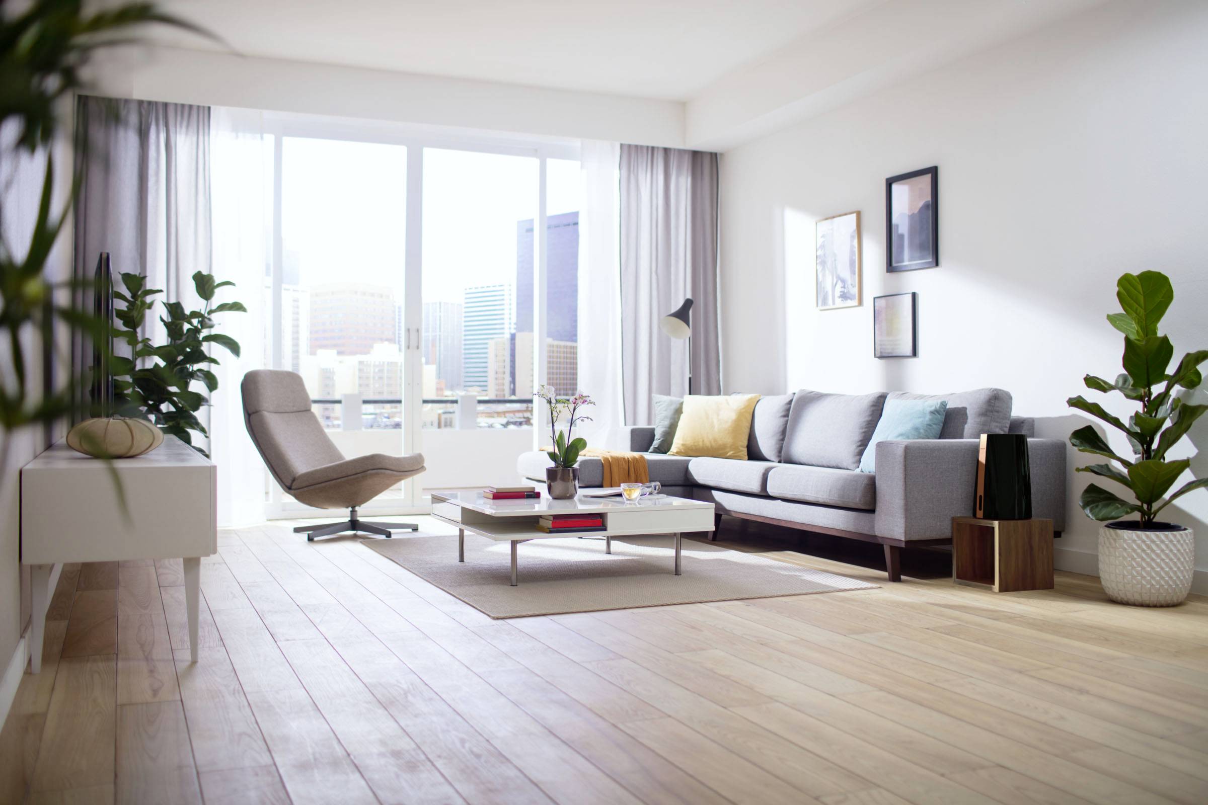 Helles stue med store vinduer, moderne sofa, lænestol, sofabord og planter. Udsigt til byens silhuet i baggrunden.