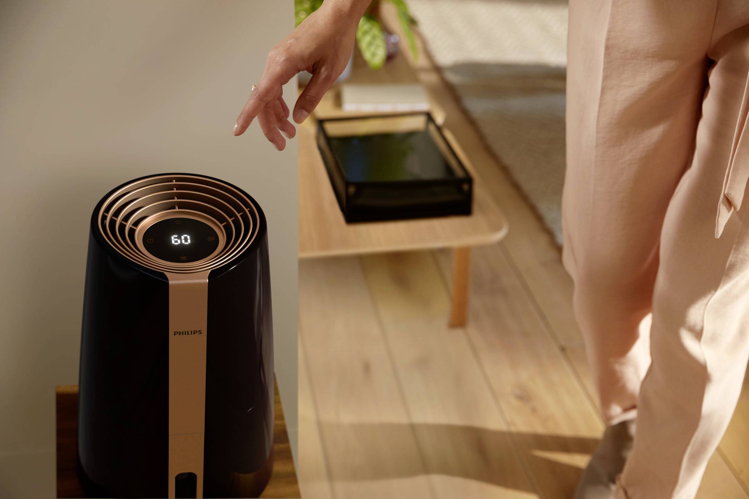 En person betjener en moderne luftrenser med digital display på et træbord i et veloplyst rum.