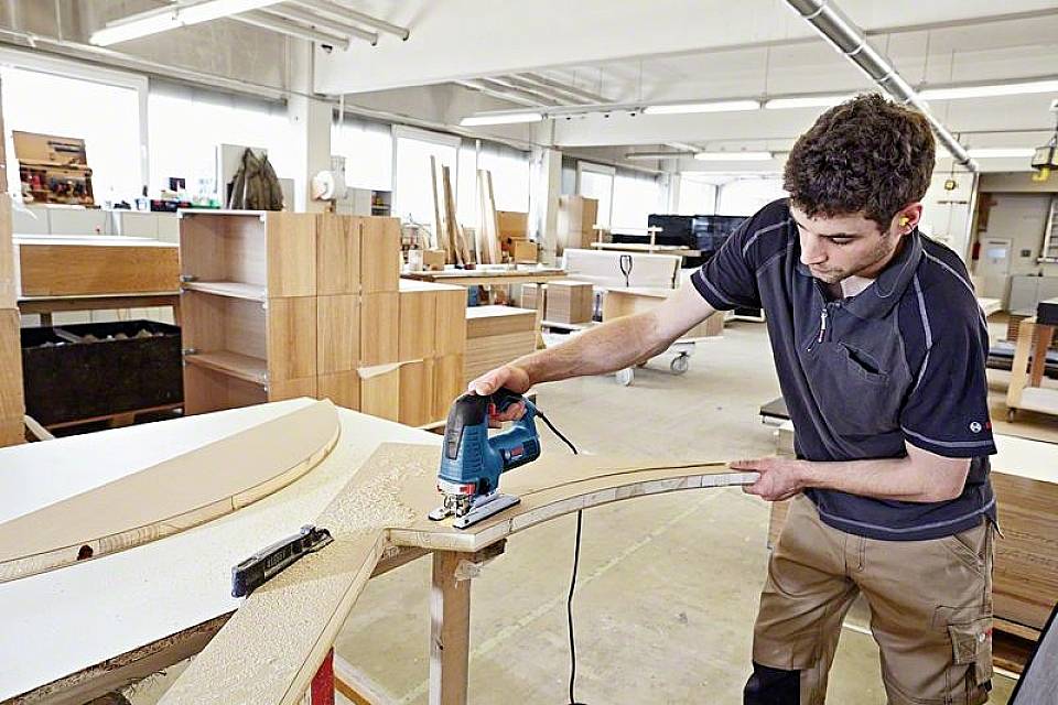 En mand i et værksted bruger en elektrisk stiksav til at skære et buet stykke træ på et bord.