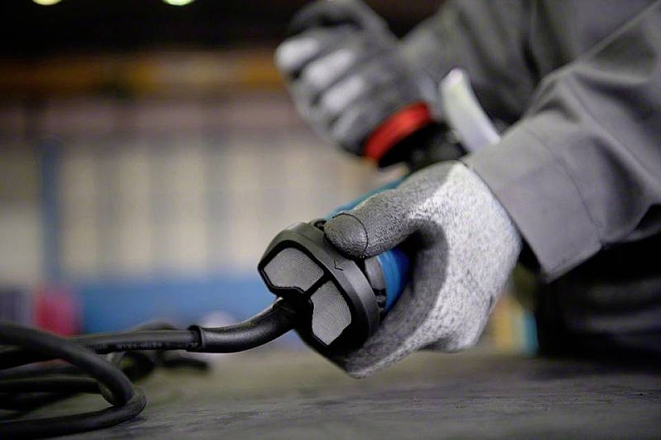 En person med grå handsker holder en elektrisk stik. Baggrunden er sløret og viser et industrielt miljø.