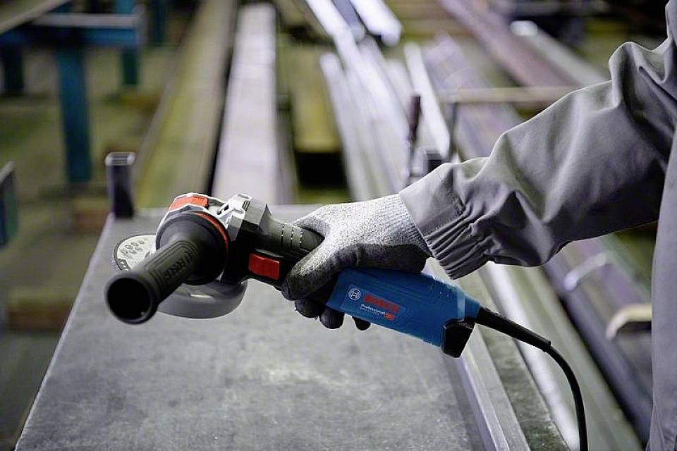 En person i arbejdstøj holder en vinkelsliber over et metalbord. Beskyttelseshandsker er synlige.