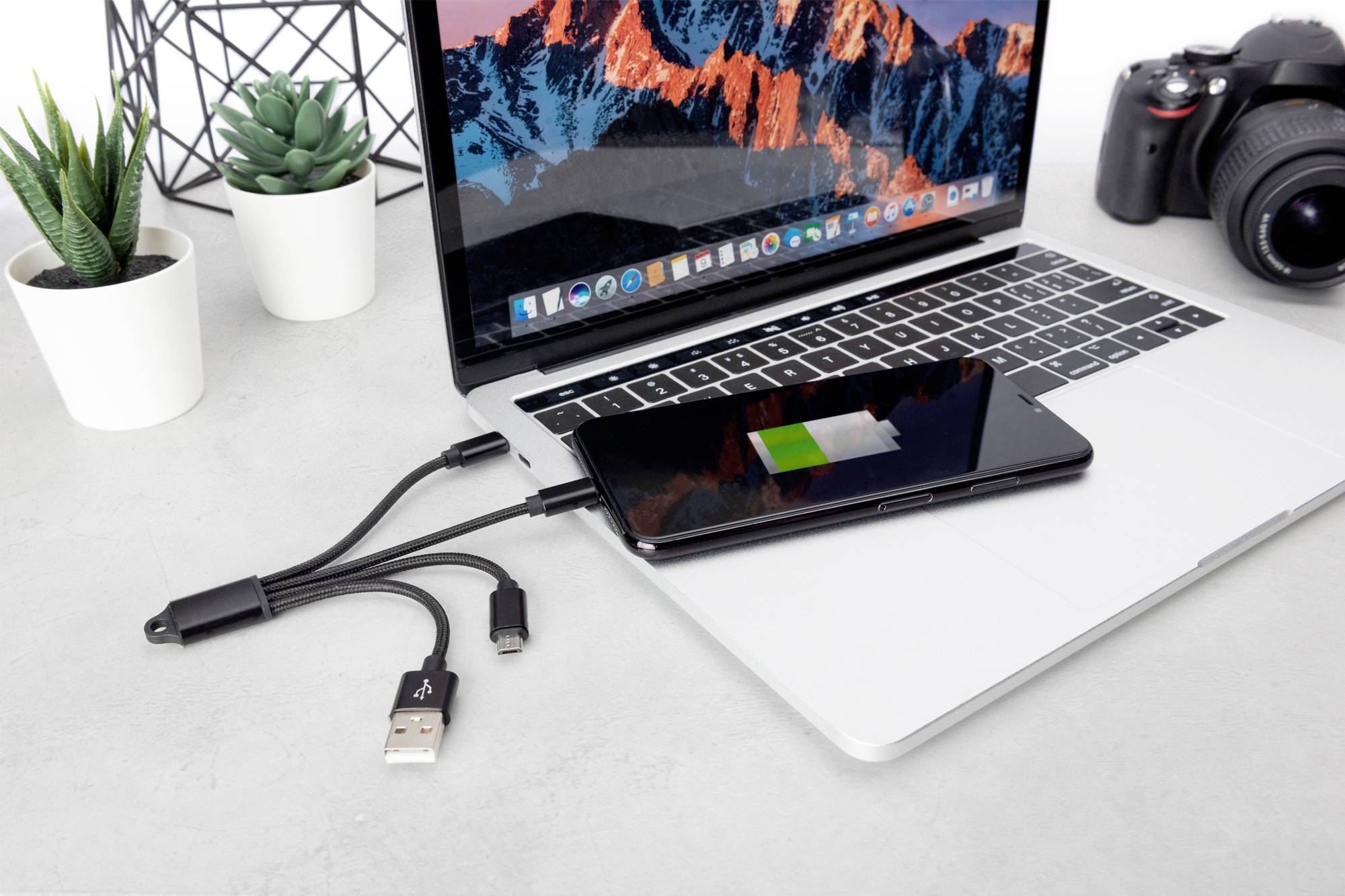 En smartphone tilsluttes og oplades via en USB-hub til en bærbar computer. I baggrunden ses to planter og et kamera.