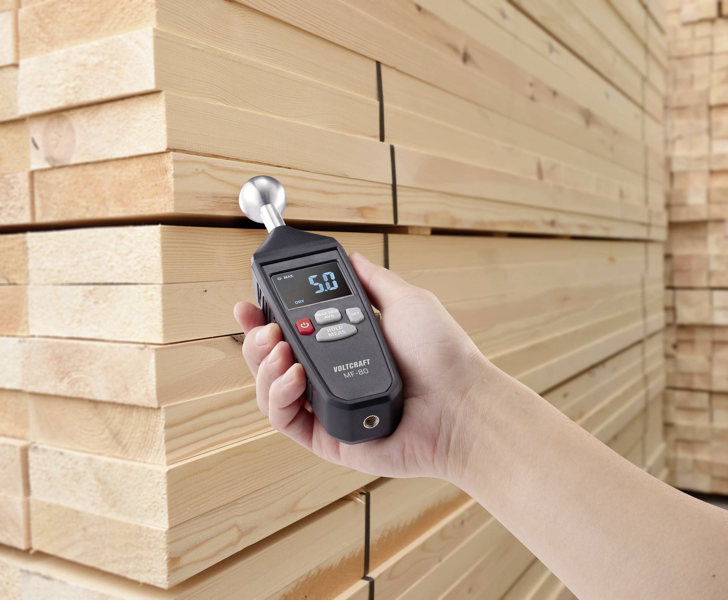 En hånd holder et fugtighedsmålingsapparat foran stablet træbrædder. Displayet viser en værdi på 5,0.