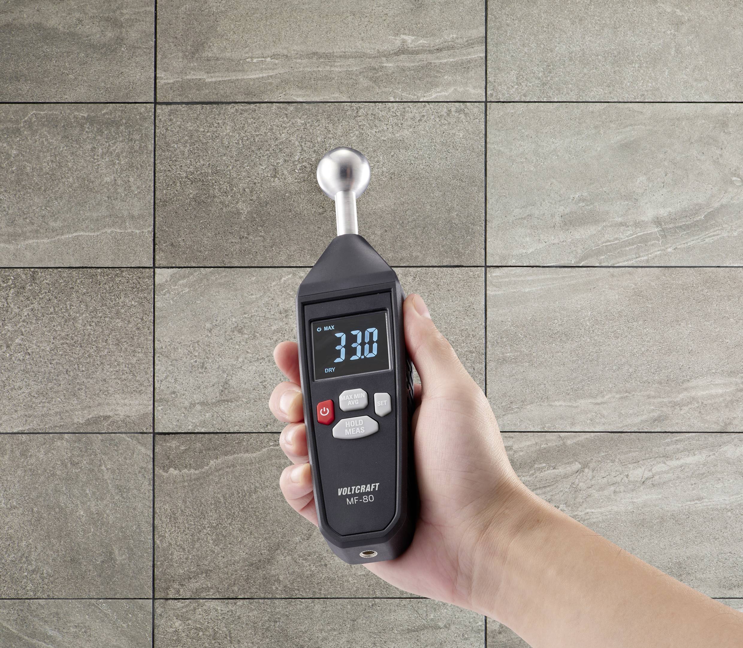 En hånd holder en digital måler foran en væg med beige fliser. Displayet viser '3.30'.