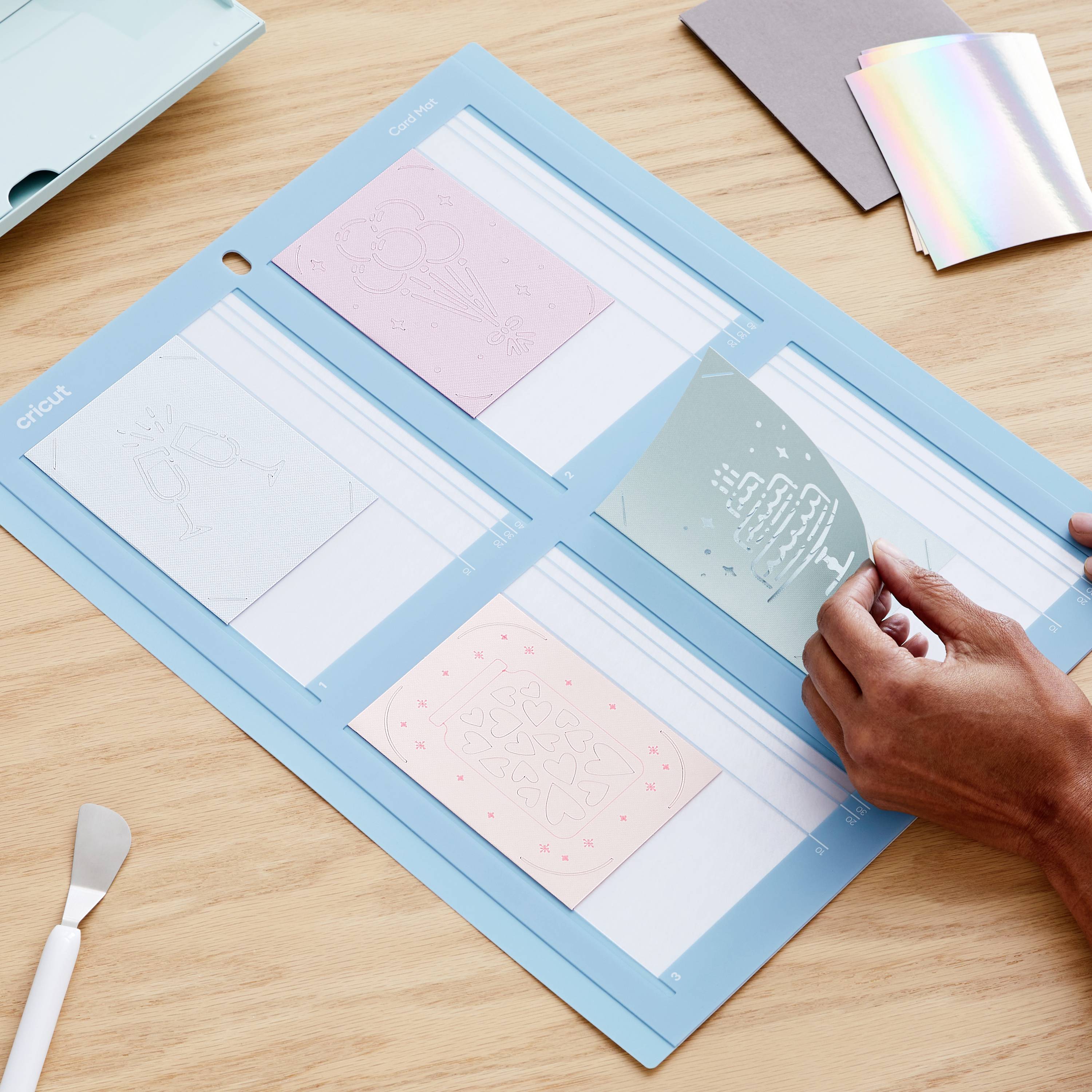 En person bruger en Cricut-maskine til at placere designs på en blå måtte. Forskellige materialer og værktøjer ligger klar.