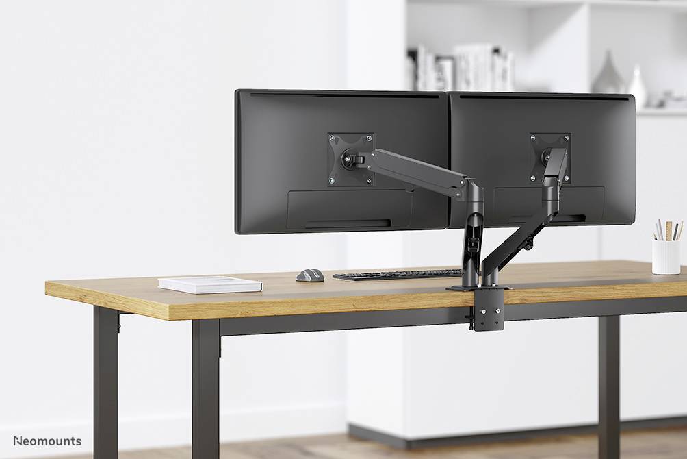 Et moderne skrivebord med to monitorer monteret ved siden af hinanden på en justerbar arm, et tastatur nedenunder og en kop til højre.