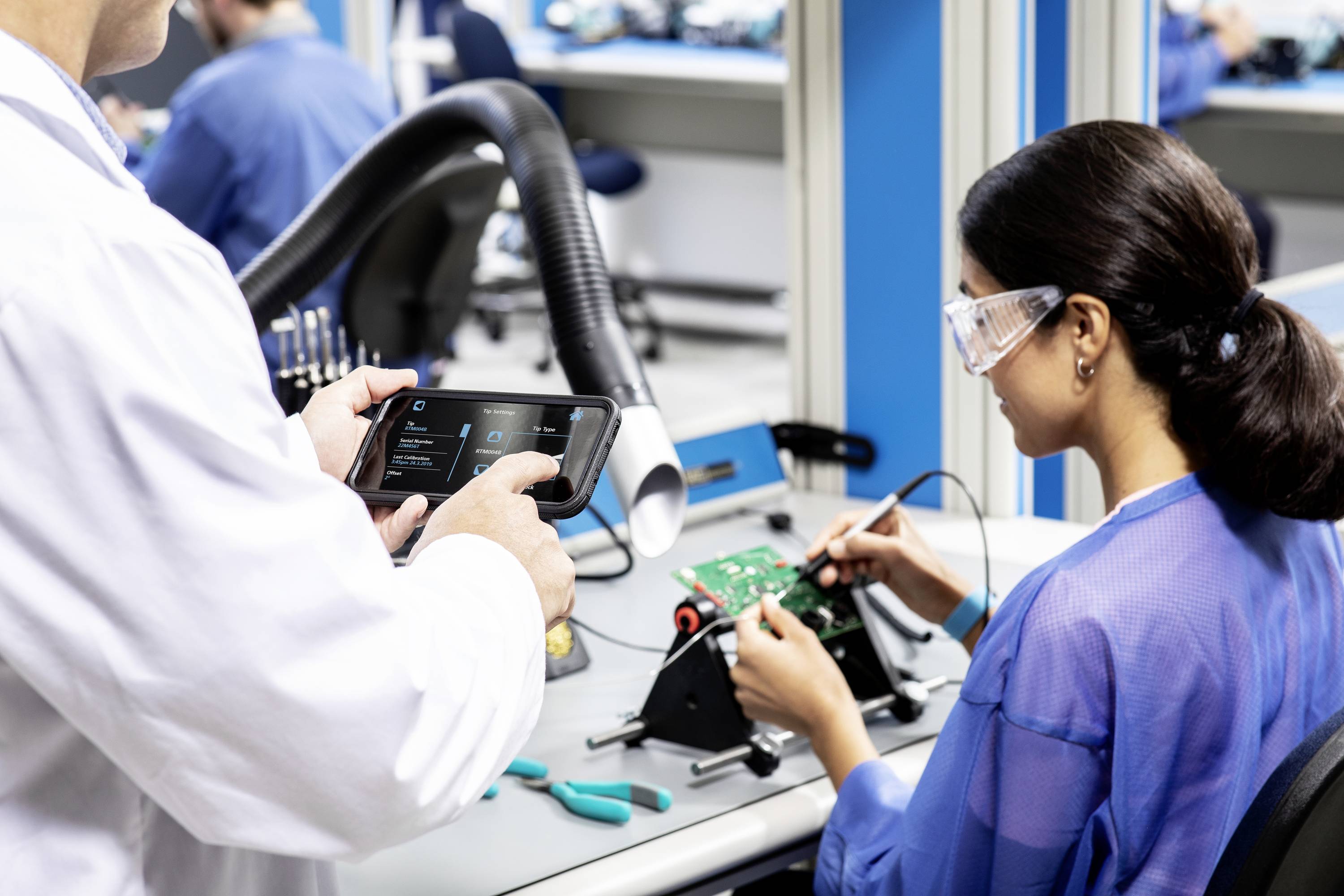 'To personer i et laboratorium. En person arbejder med elektroniske komponenter, mens den anden bruger et tablet til målinger.'