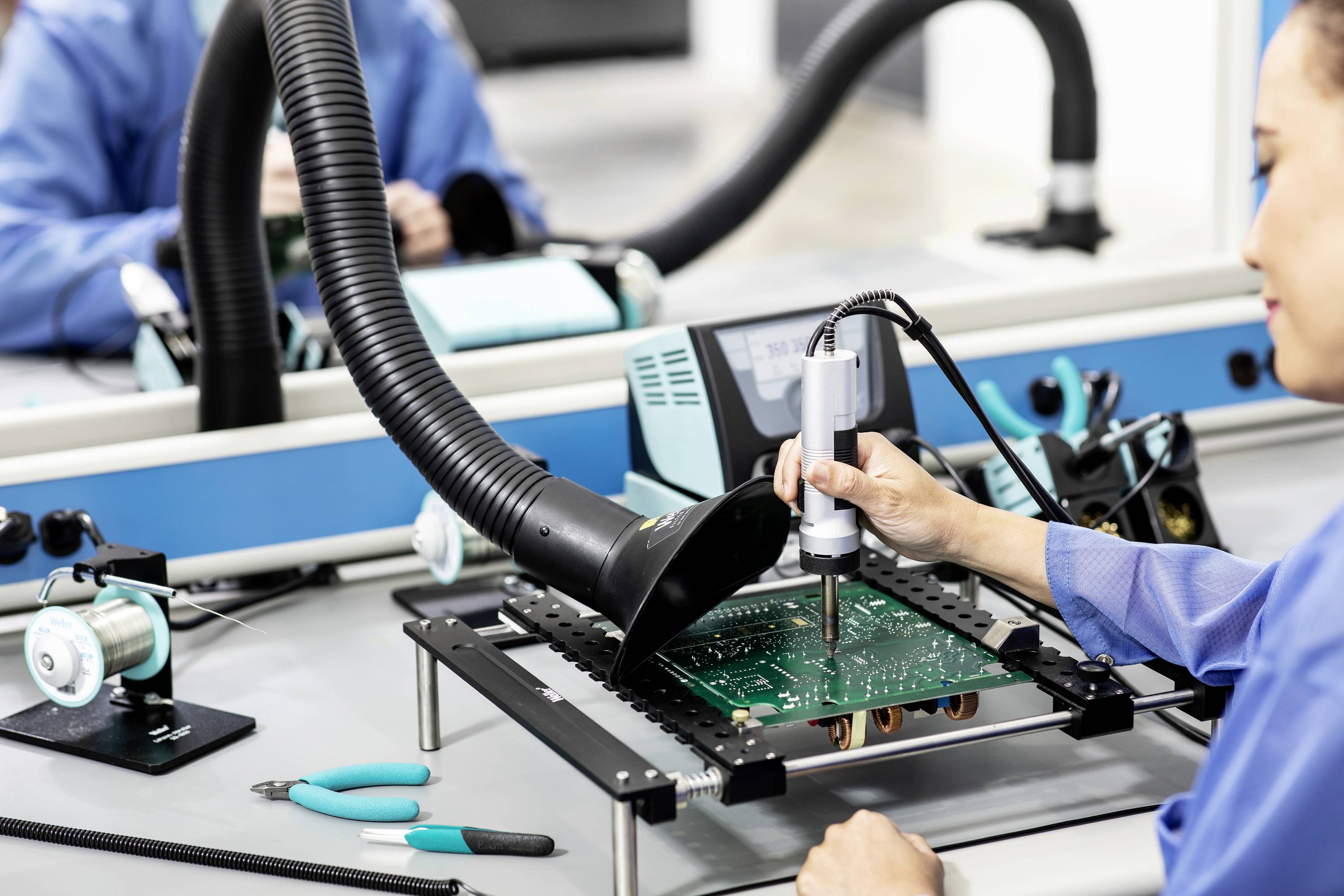 En ingeniør lodder et printpanel i et elektronikværksted, omgivet af teknisk udstyr og værktøjer som tænger og loddekolber.