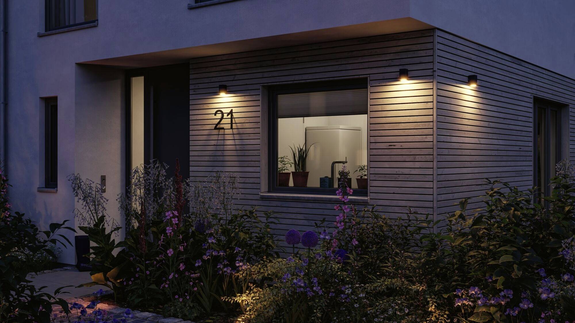 Et moderne hus ved tusmørke, oplyst af udendørslamper. En vindueskarm med planter er synlig. Husnummeret '21' kan ses.