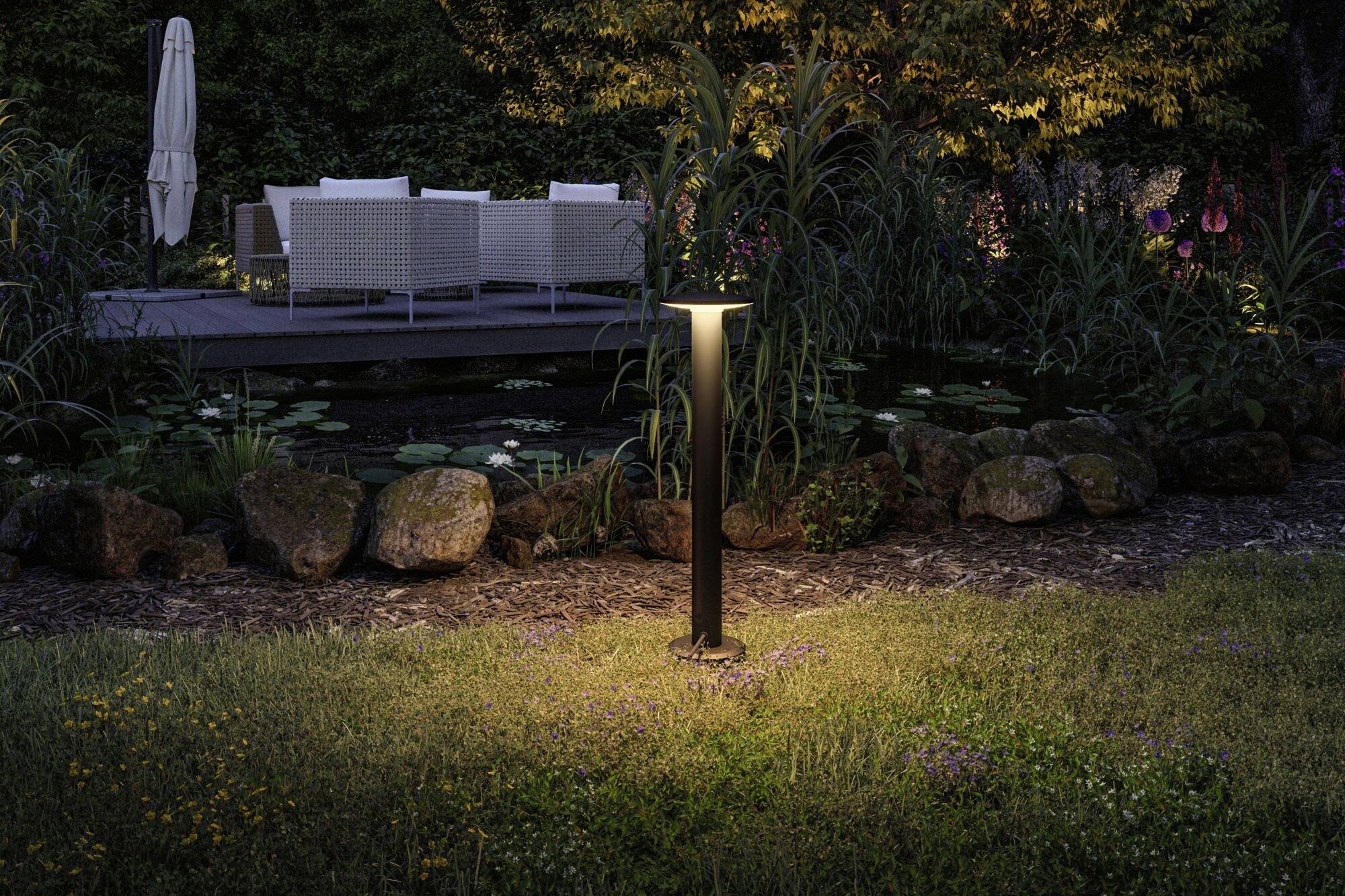 Oplyst have med dam, klipper og græsser; i baggrunden havemøbler og træer, der skaber en hyggelig aftenstemning.