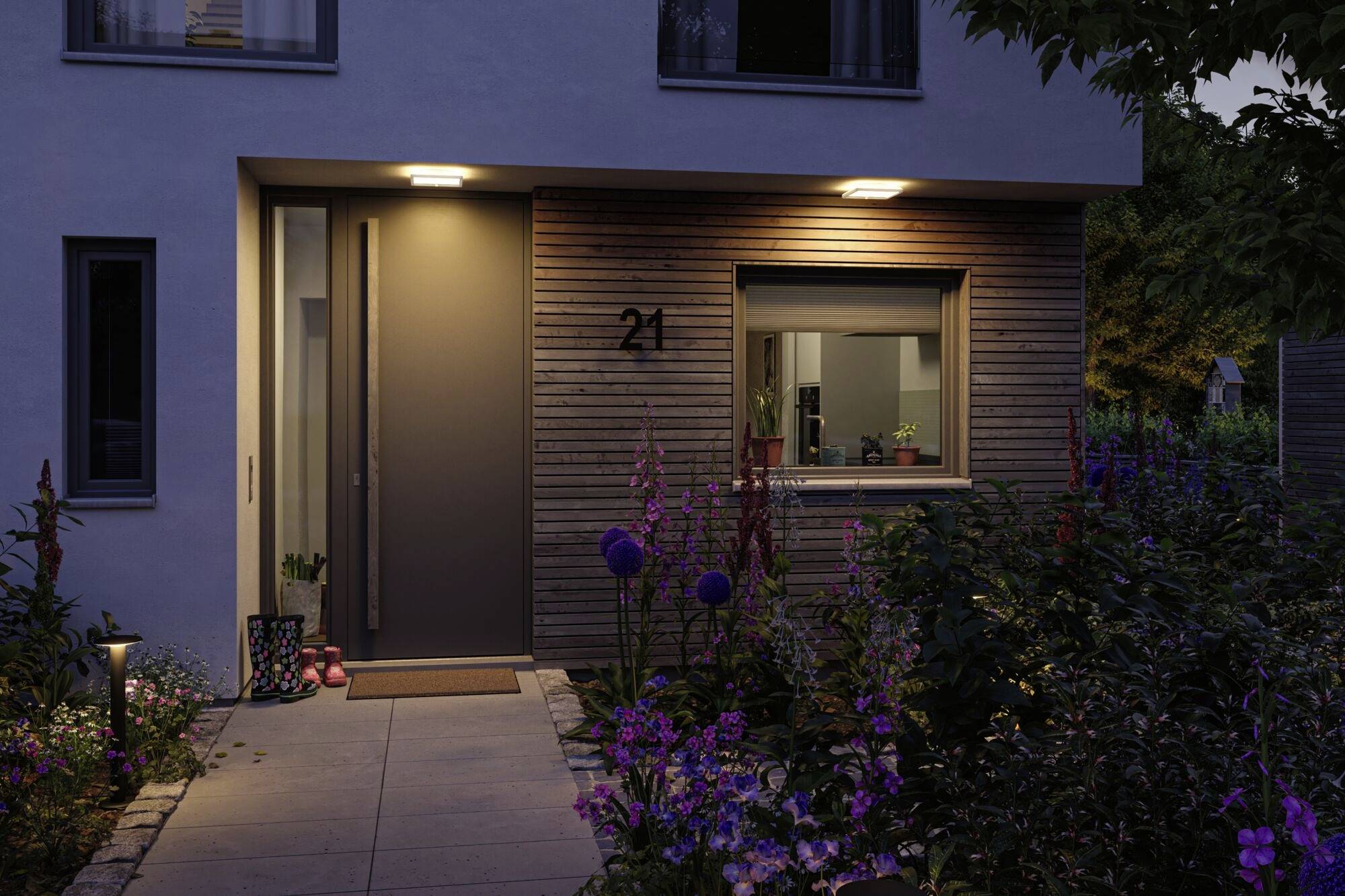 Et moderne hus med oplyst indgang. Foran huset blomstrer blomster. I vinduet står stueplanter. Husnummer 21 synligt.