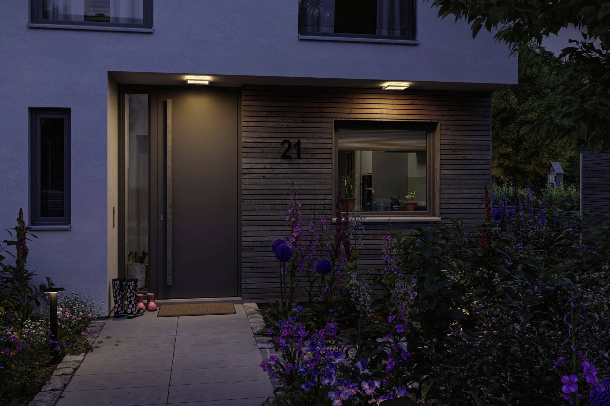 Hoveddør til et moderne hus ved tusmørke, omgivet af blomsterhave, husnummer 21 synligt. Lamper oplyser indgangen.