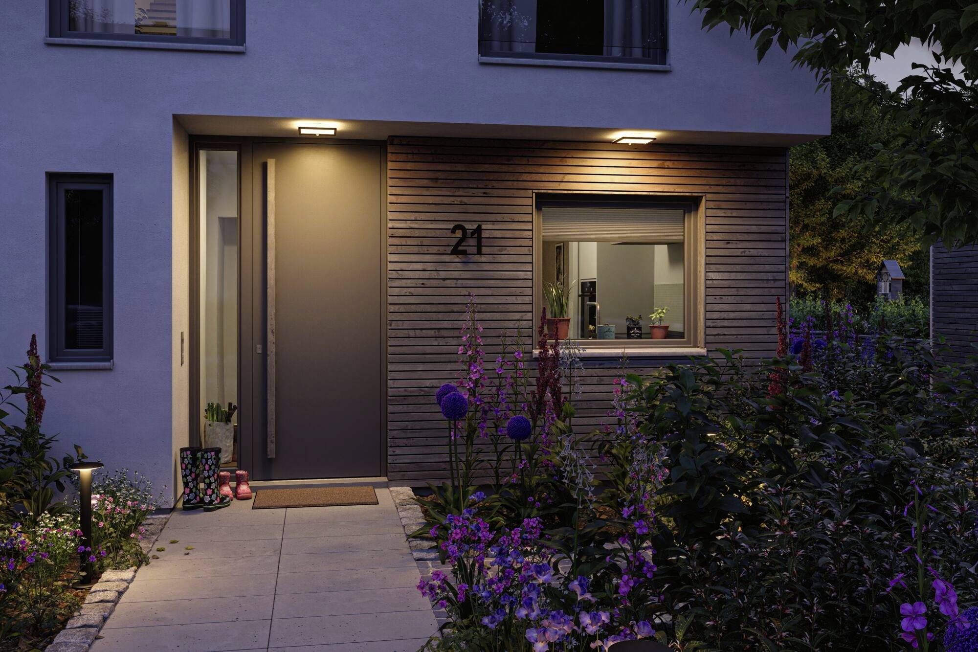 Indgangsparti til et moderne hus ved tusmørke, omgivet af blomstrende blomster. Oplyst husnummer 21, vindue viser planter indvendigt.