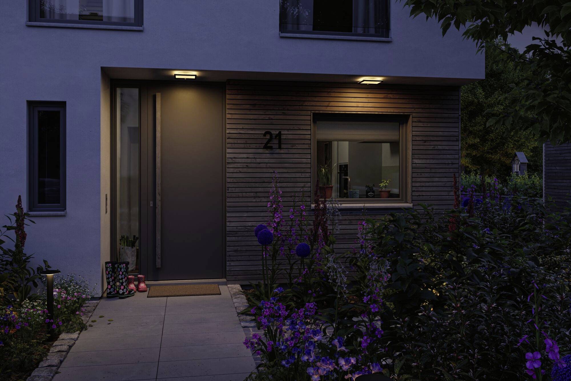 Indgang til et moderne hus ved tusmørke, oplyst af to lamper. Omgivet af blomstrende planter og en velplejet have.