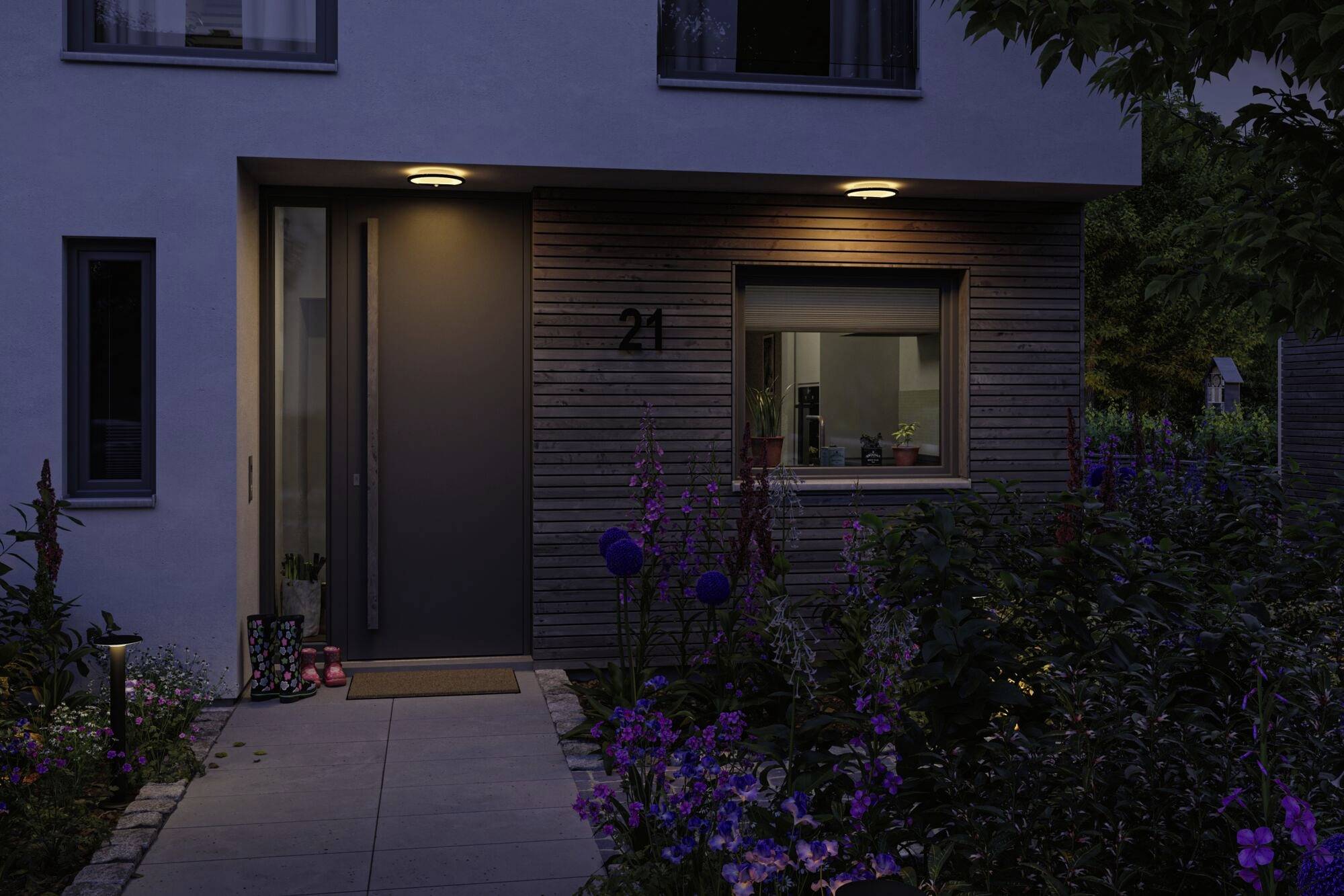 Et moderne hus om natten med oplyst indgang. Et vindue med udsigt ind i det indre viser planter. Blomster omkranser vejen til døren.