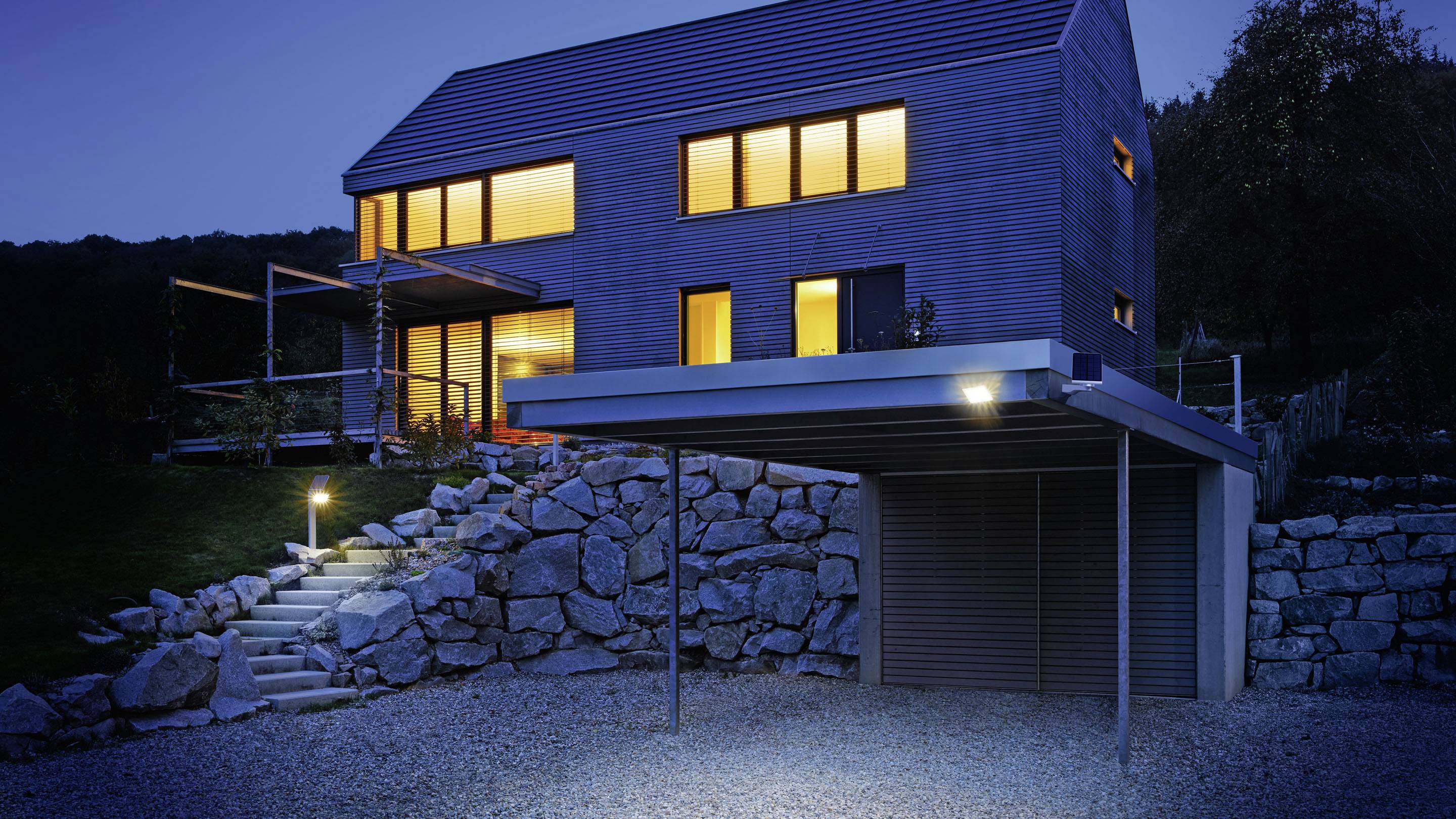 Et moderne hus med store, oplyste vinduer om natten, omgivet af en have og stenmure, med carport i forgrunden.
