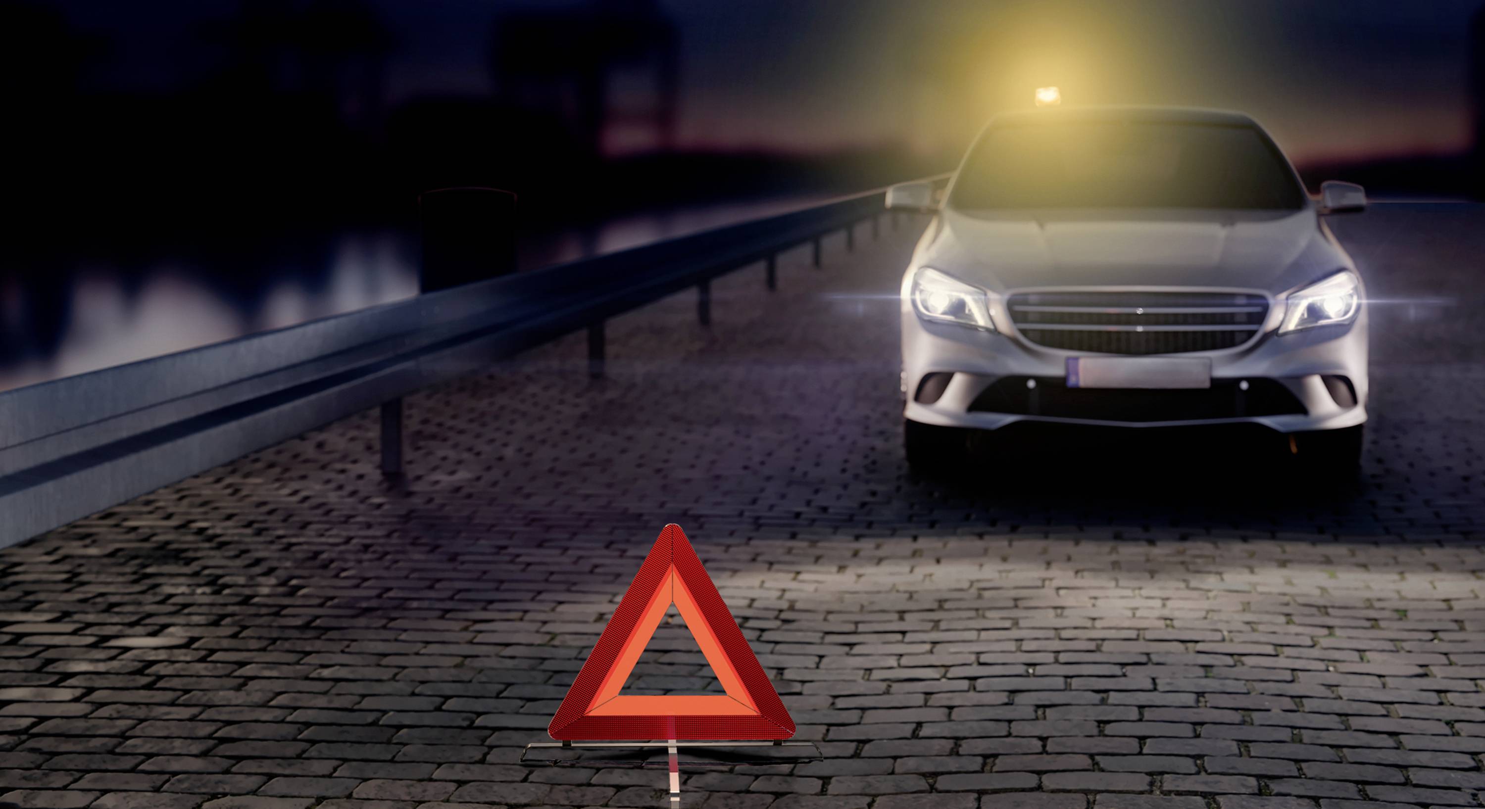 En sølvfarvet bil holder om natten på en brolagt vej med tændte advarselsblink. Et rødt advarselstrekant er placeret foran bilen.