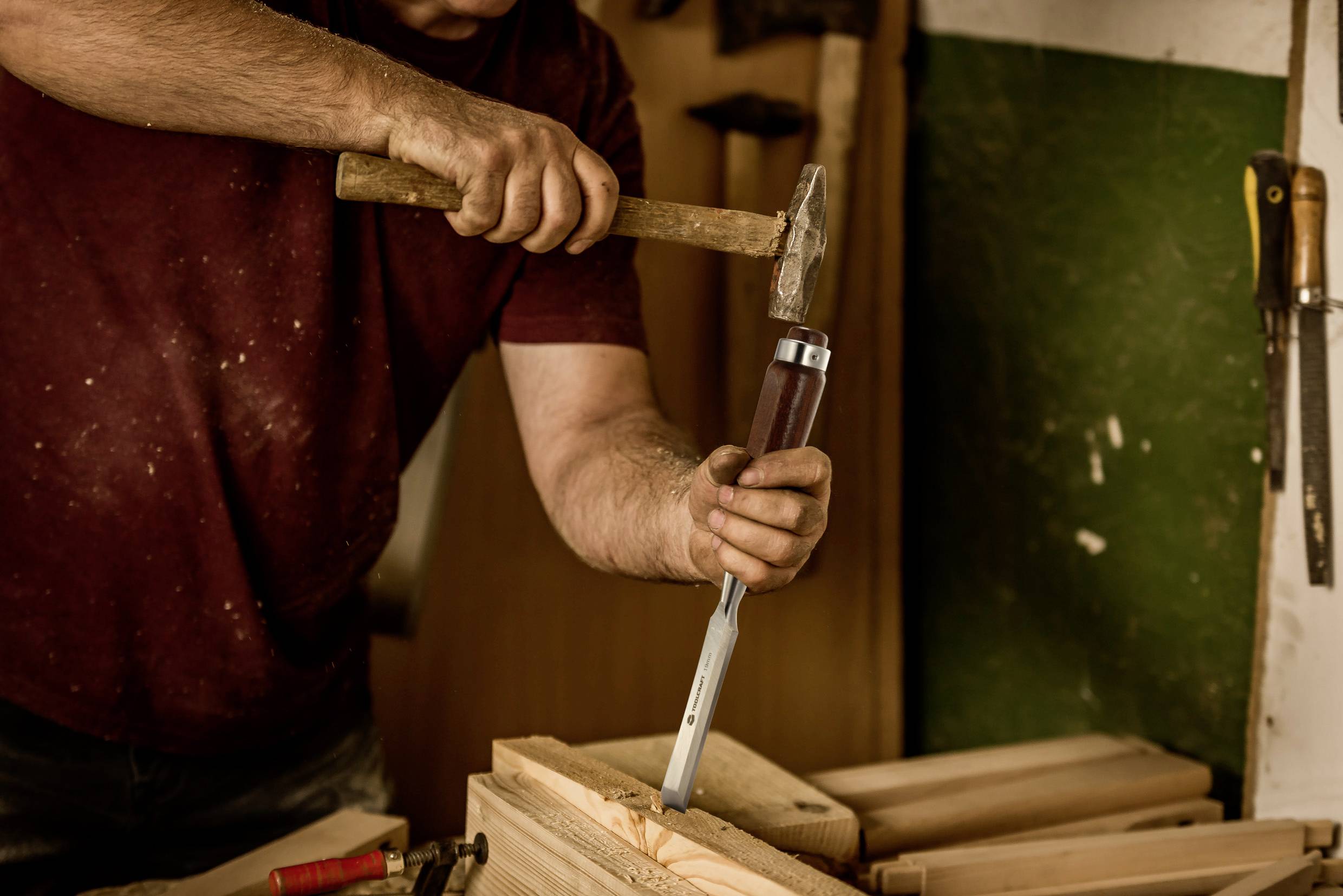 En person bearbejder et stykke træ med hammer og mejsel i et værksted, omgivet af værktøj på væggen.