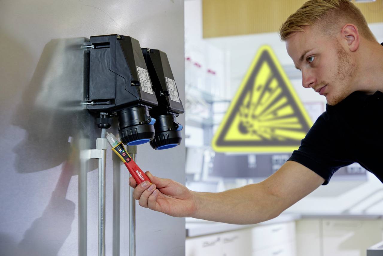 En mand kontrollerer elektriske apparater med en testanordning. I baggrunden et skilt med advarsel om eksplosionsfare.